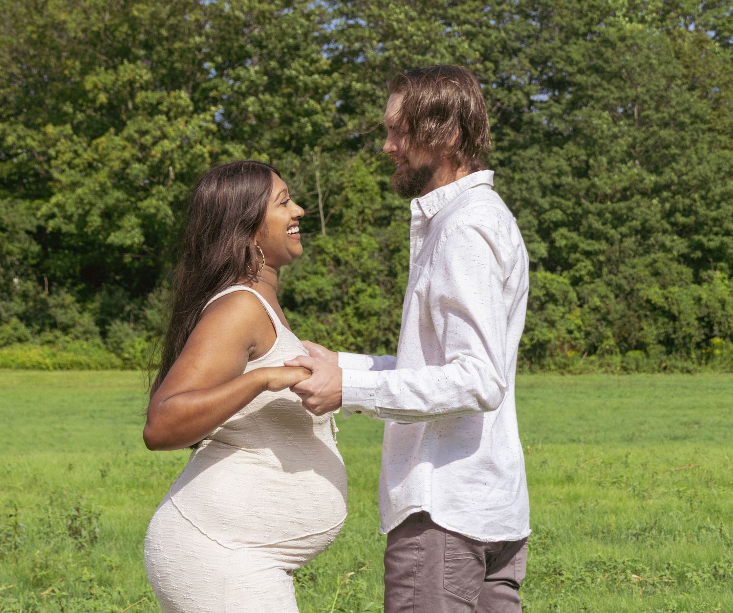 Pregnant woman in white dress holds hands with man in green field, sharing a joyful moment outdoors.