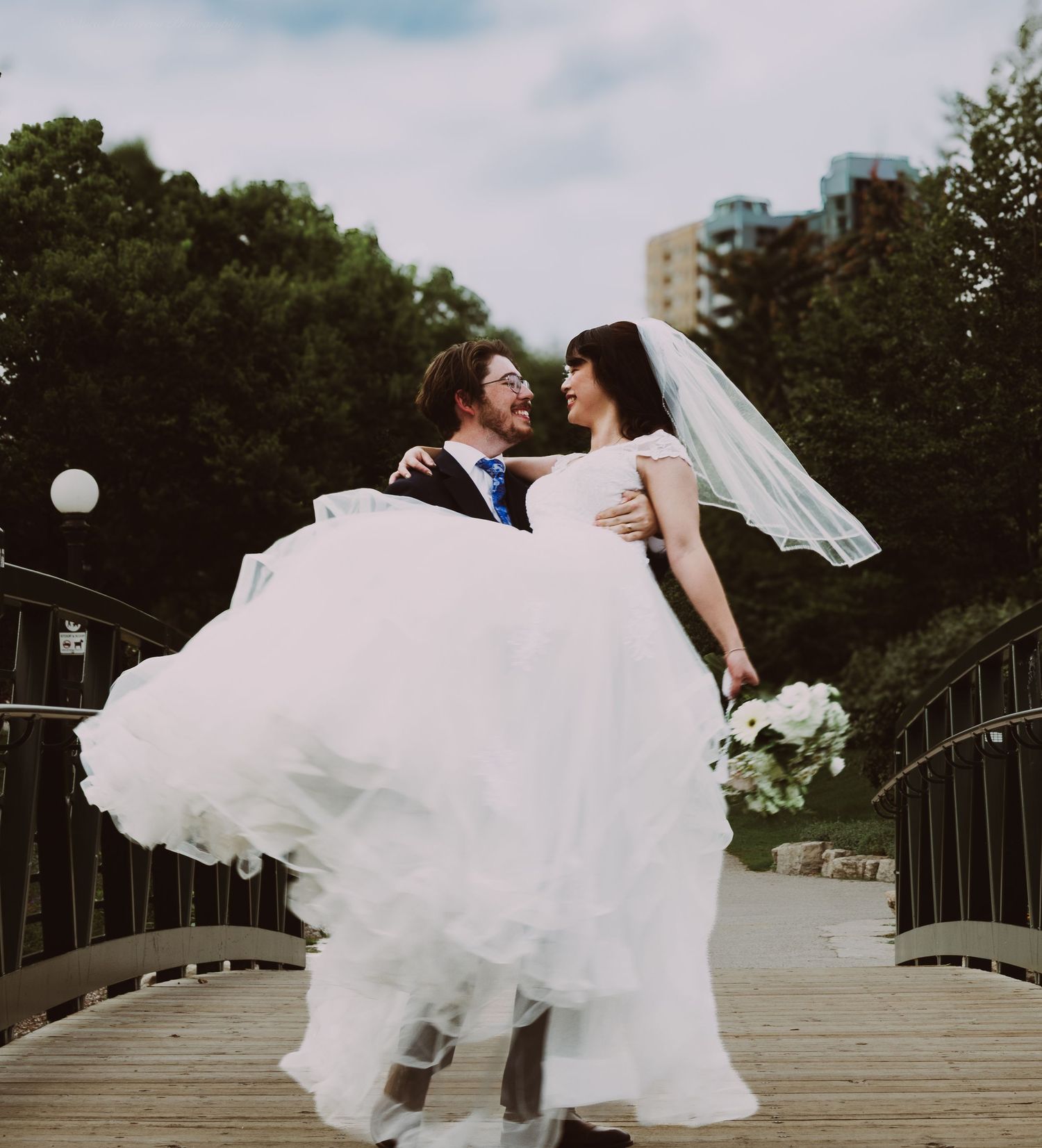 Groom lifting bride in white wedding gown and veil on a bridge surrounded by lush green trees.