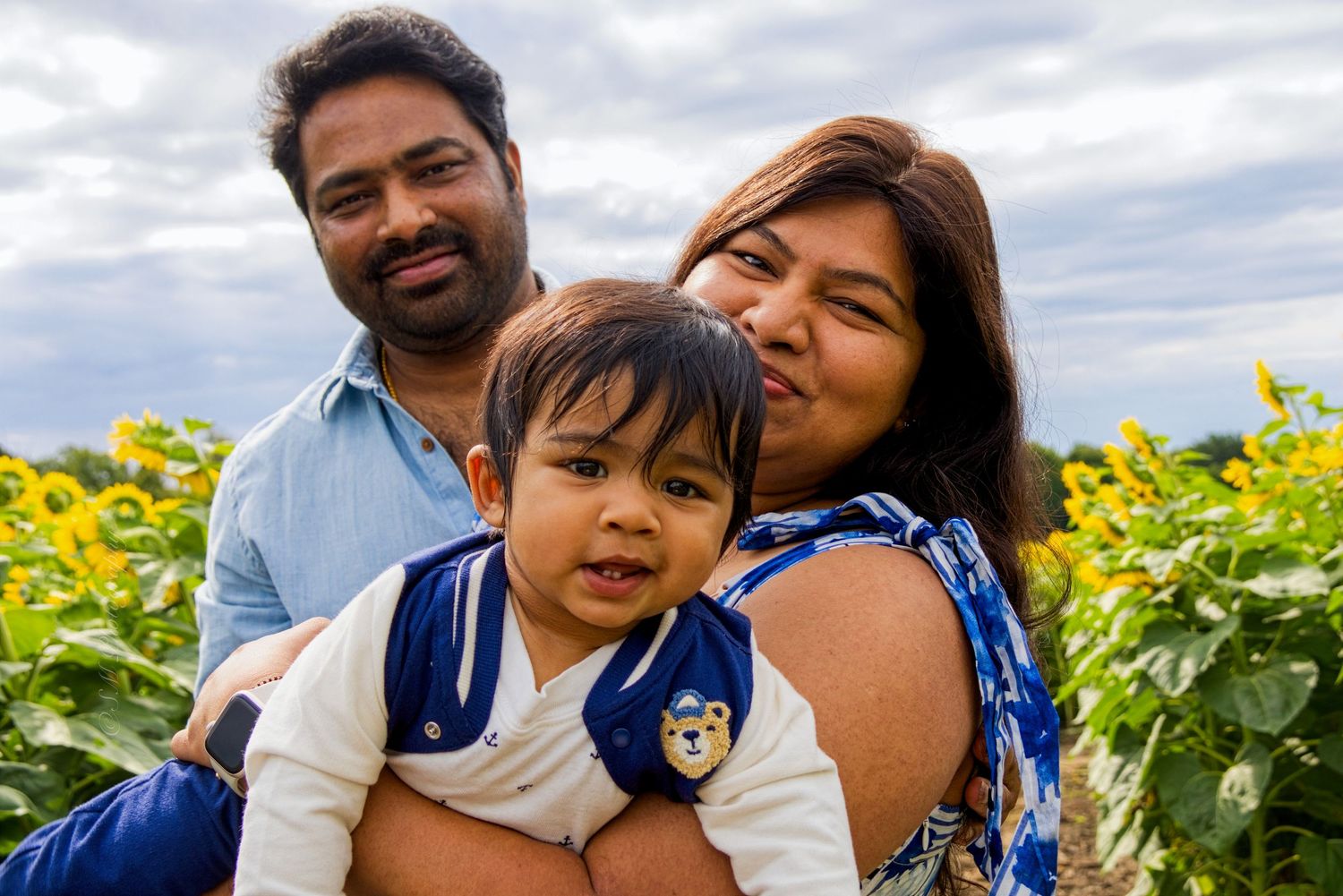 A happy family of three poses together with their baby in a sunflower field.