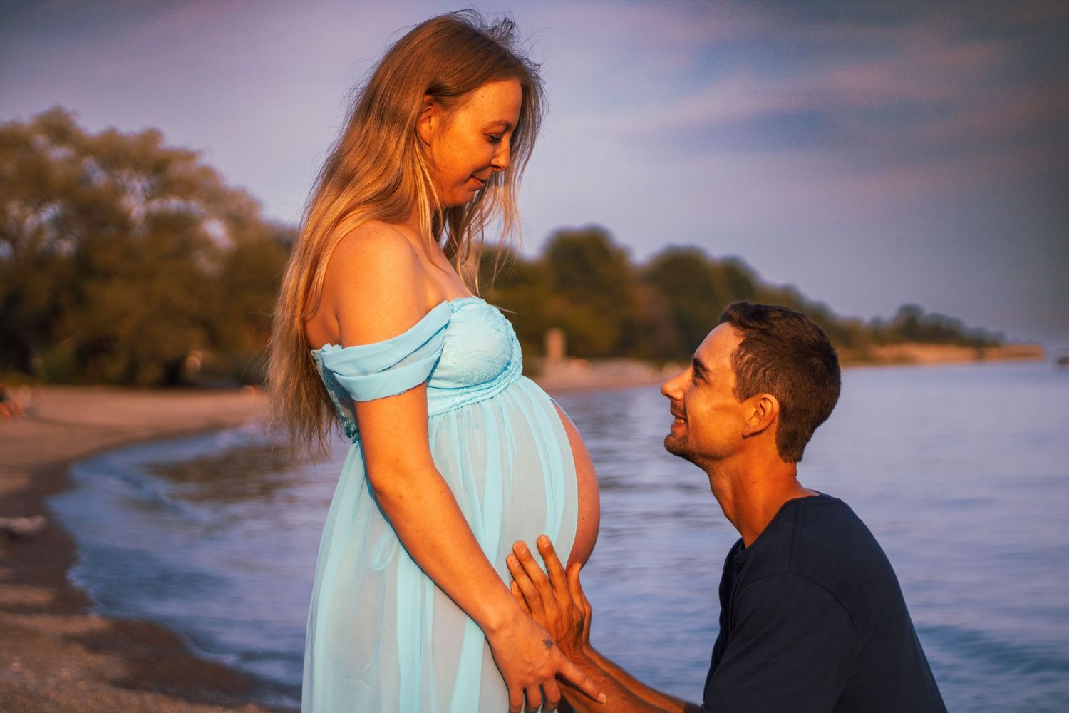 Pregnant woman in teal dress on beach with partner kneeling and touching her baby bump at sunset.