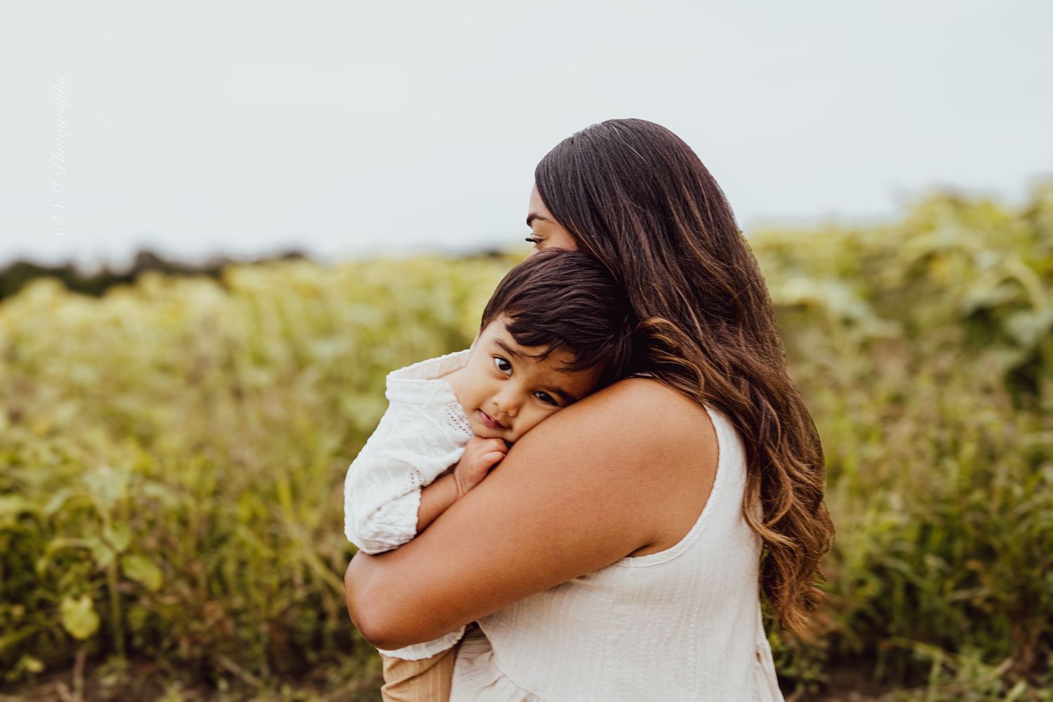 Mother with long brown hair tenderly embracing her baby in a green sunflower field on an overcast day.