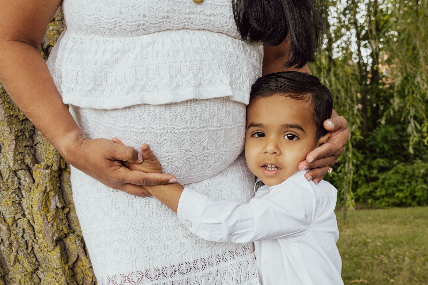 A young child in white hugs a pregnant woman's belly outdoors near a tree during a maternity photo session.