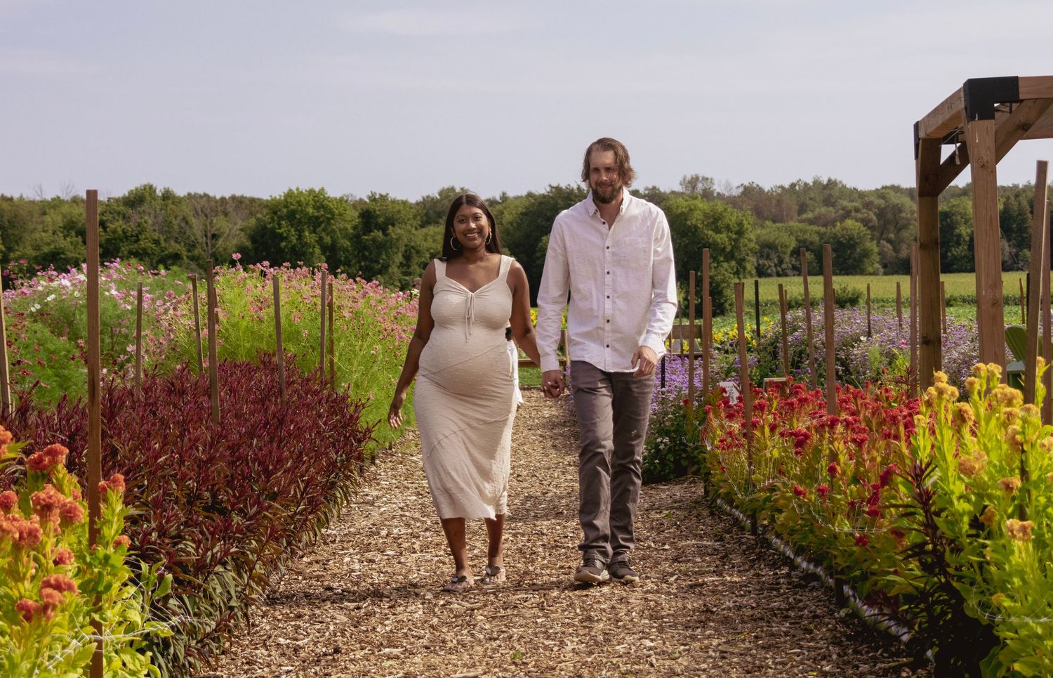 Pregnant woman in white dress walks with partner through colorful flower garden path on sunny day.