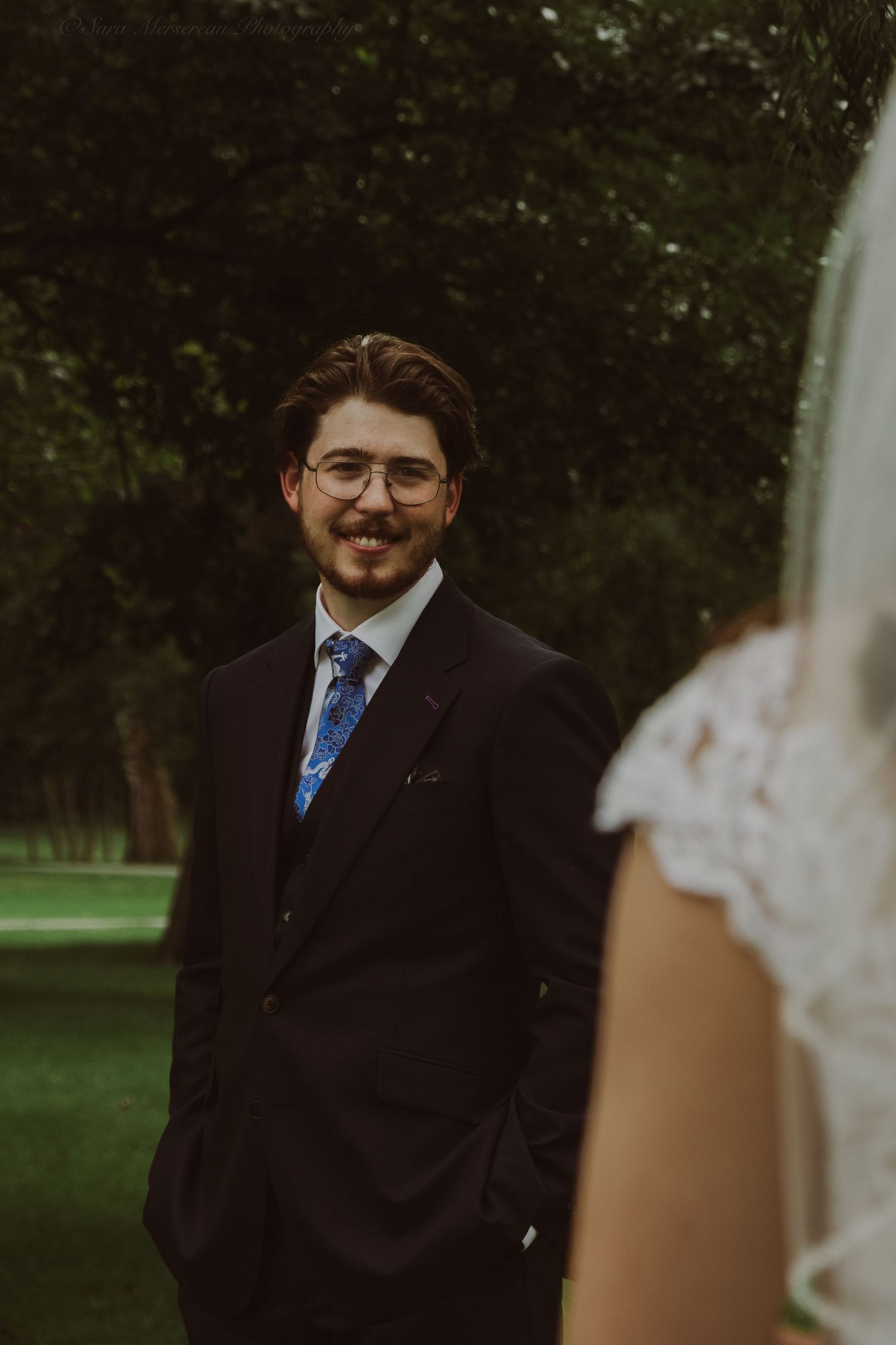 Groom in black suit and blue floral tie smiling at bride during outdoor wedding ceremony.
