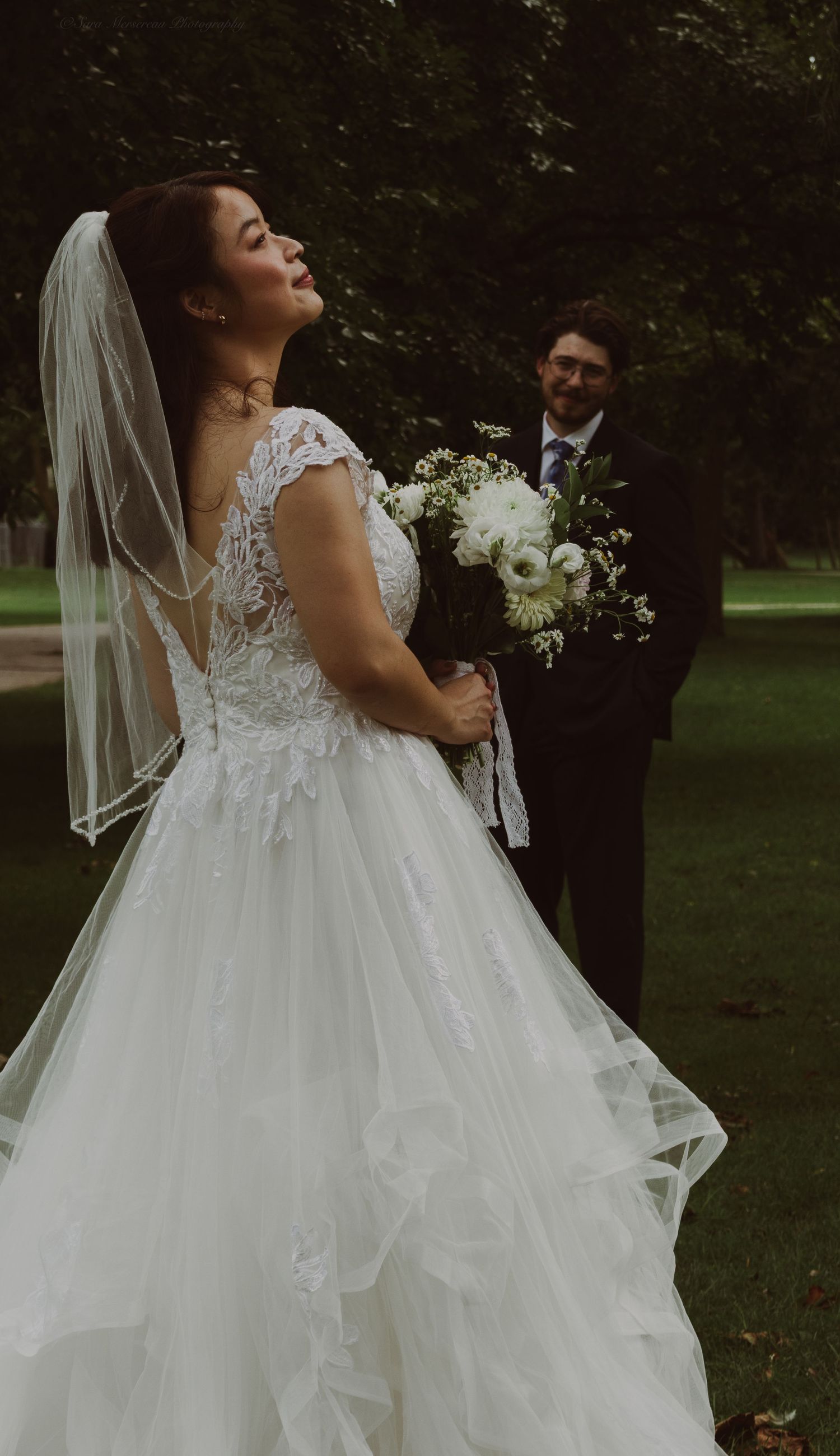 Bride in white lace ballgown and veil holds bouquet while groom in black suit stands behind her outdoors.