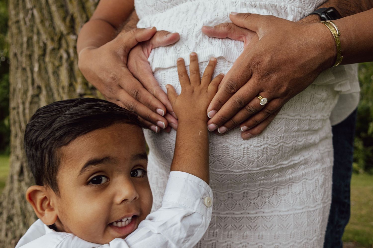 A young boy smiles while touching a pregnant woman's belly outdoors, with two adults placing hands on the bump.