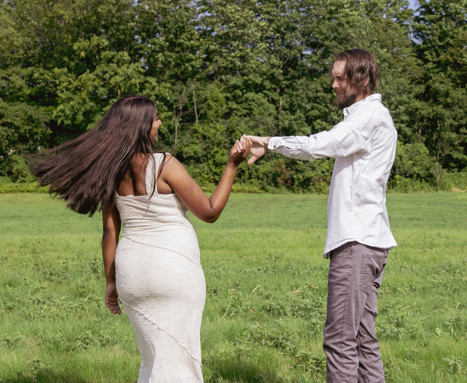 Couple dancing in a green meadow, woman in white dress with flowing hair, man in white shirt holding hands outdoors.