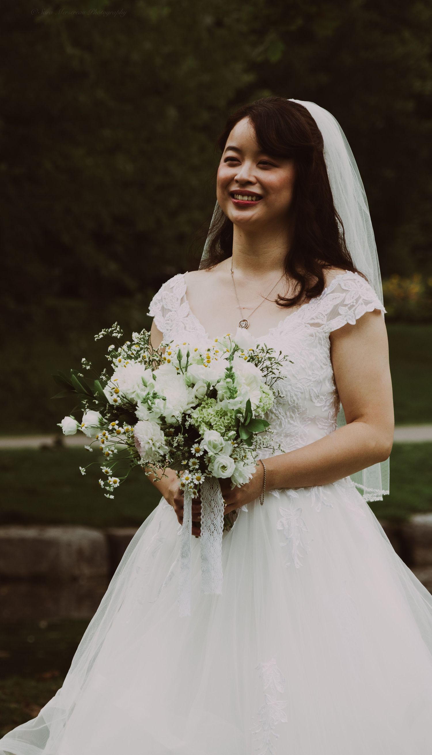 Smiling Asian bride in white lace gown and veil holding white floral bouquet outdoors.