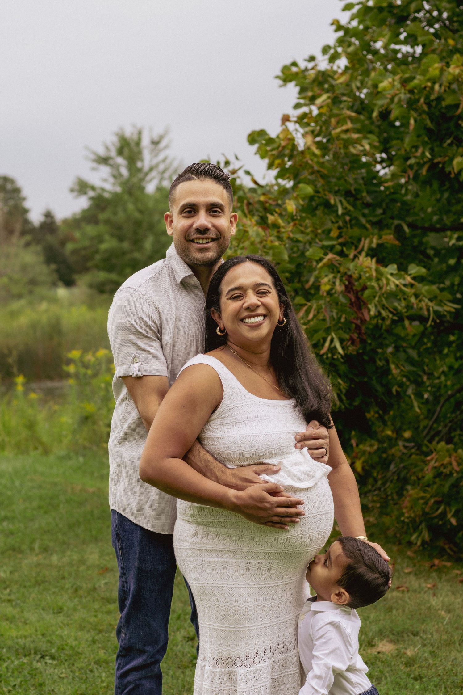 Smiling pregnant woman in white dress poses outdoors with her husband and young child in a lush green park setting.