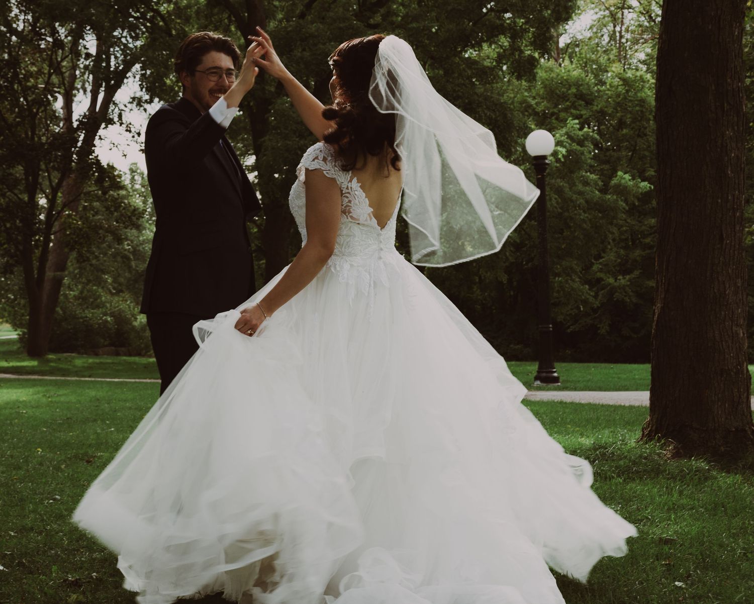A bride in a white ball gown and veil dances with a groom in a black suit outdoors on a lush green lawn.