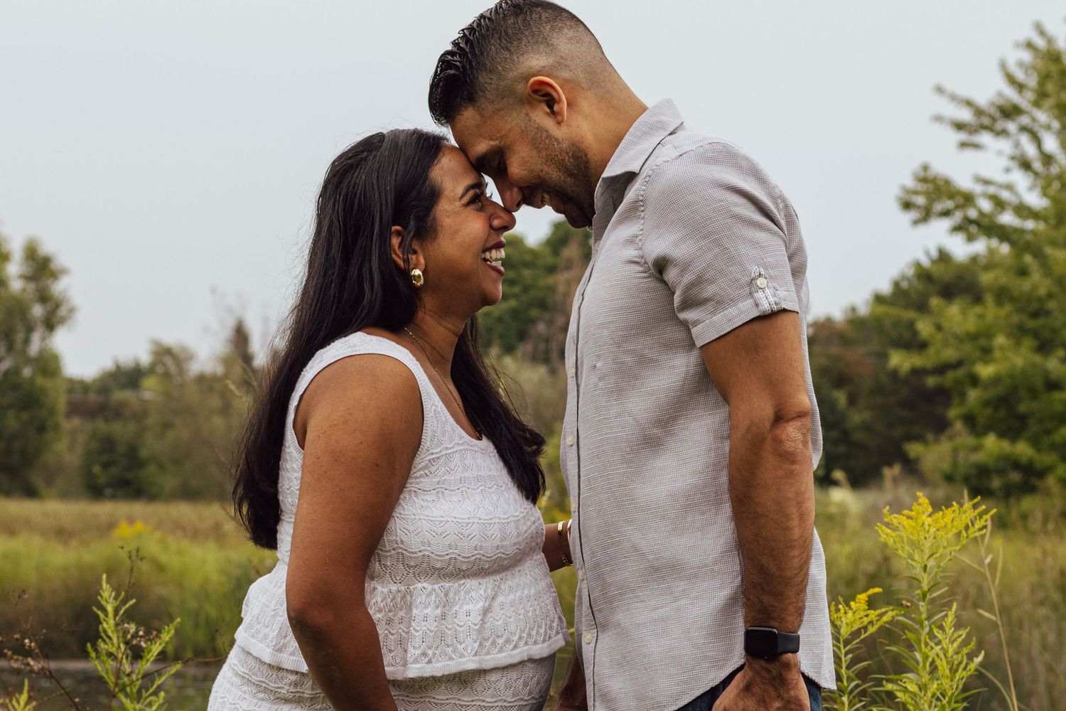 Happy couple nearly kissing while touching foreheads in a natural outdoor setting with green trees behind them.