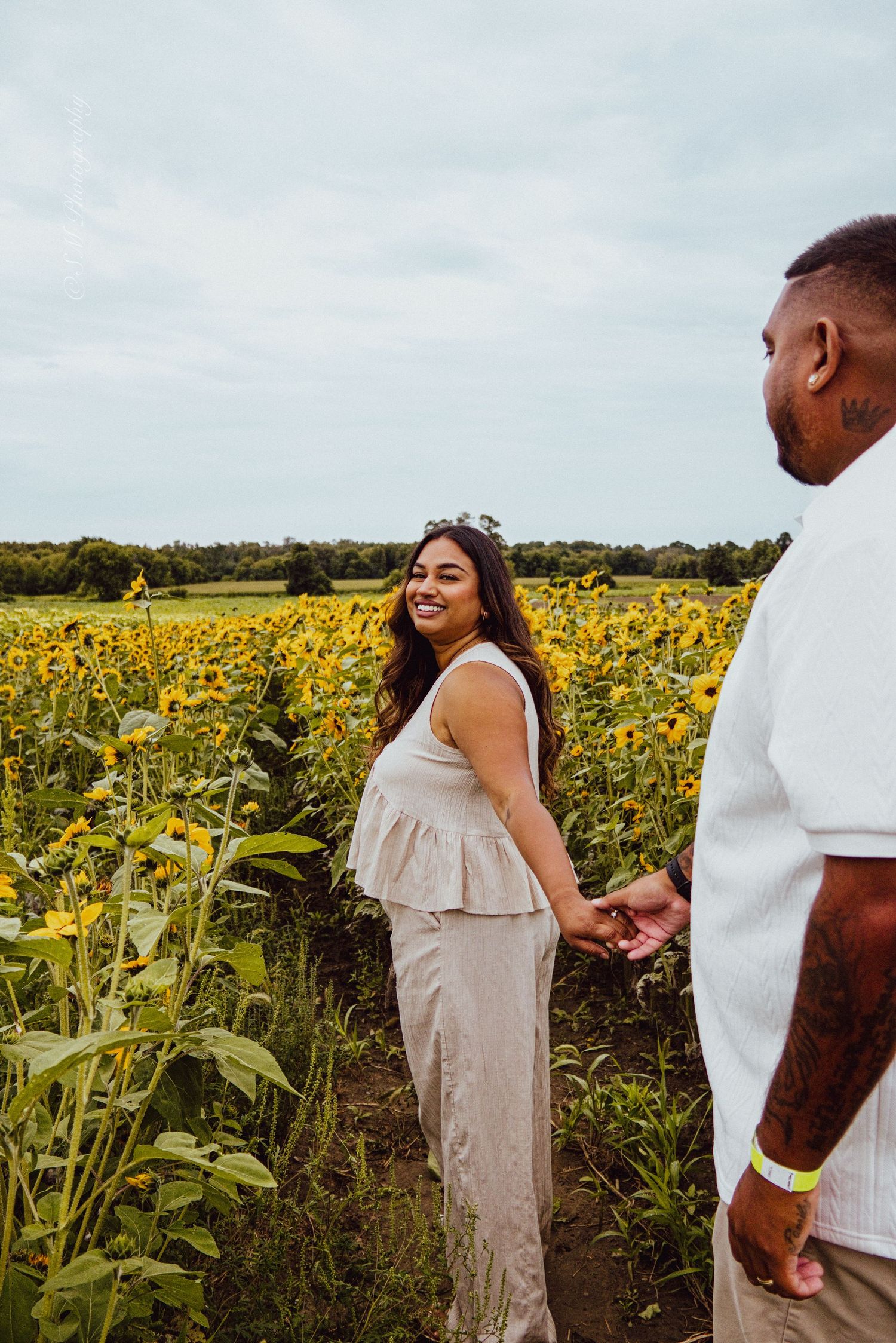 Smiling woman holding hands with her partner while walking through a bright yellow sunflower field.