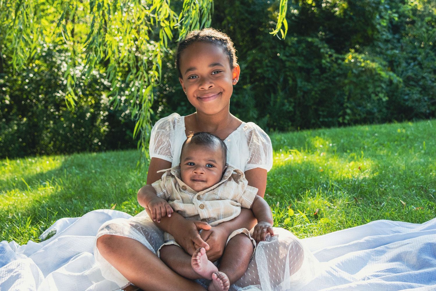 A smiling young girl sits on a blanket outdoors holding a happy baby sibling in her lap.