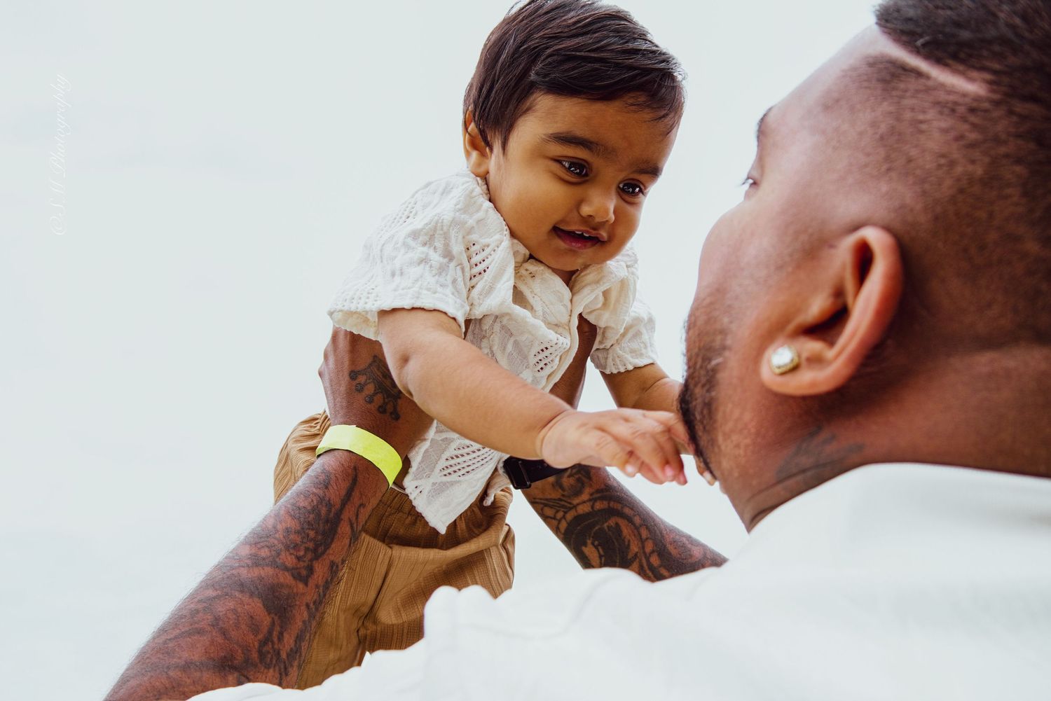 Tattooed father lovingly lifting smiling baby wearing a yellow hospital bracelet against a white background.