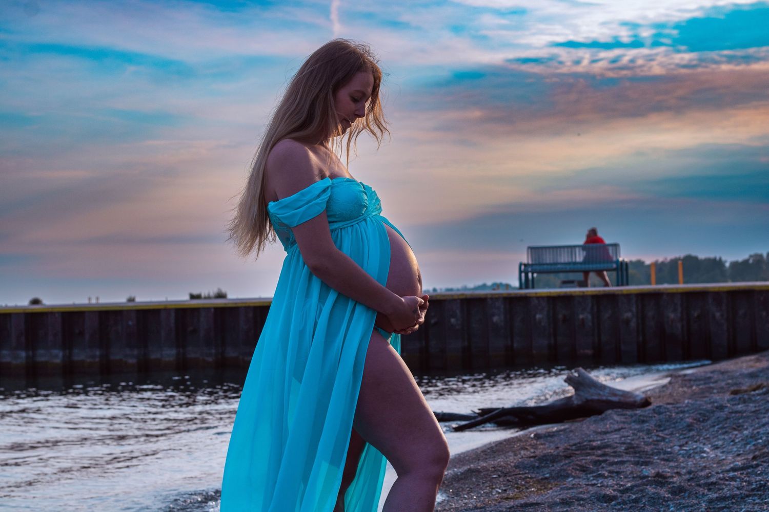 Pregnant woman in teal dress holding belly on beach at sunset.