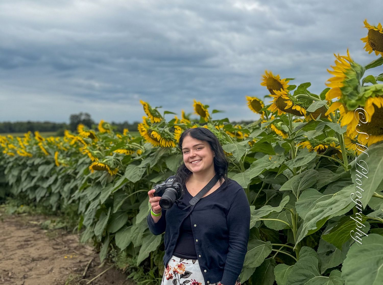 A smiling female photographer holds her camera while standing beside tall sunflowers in a field.