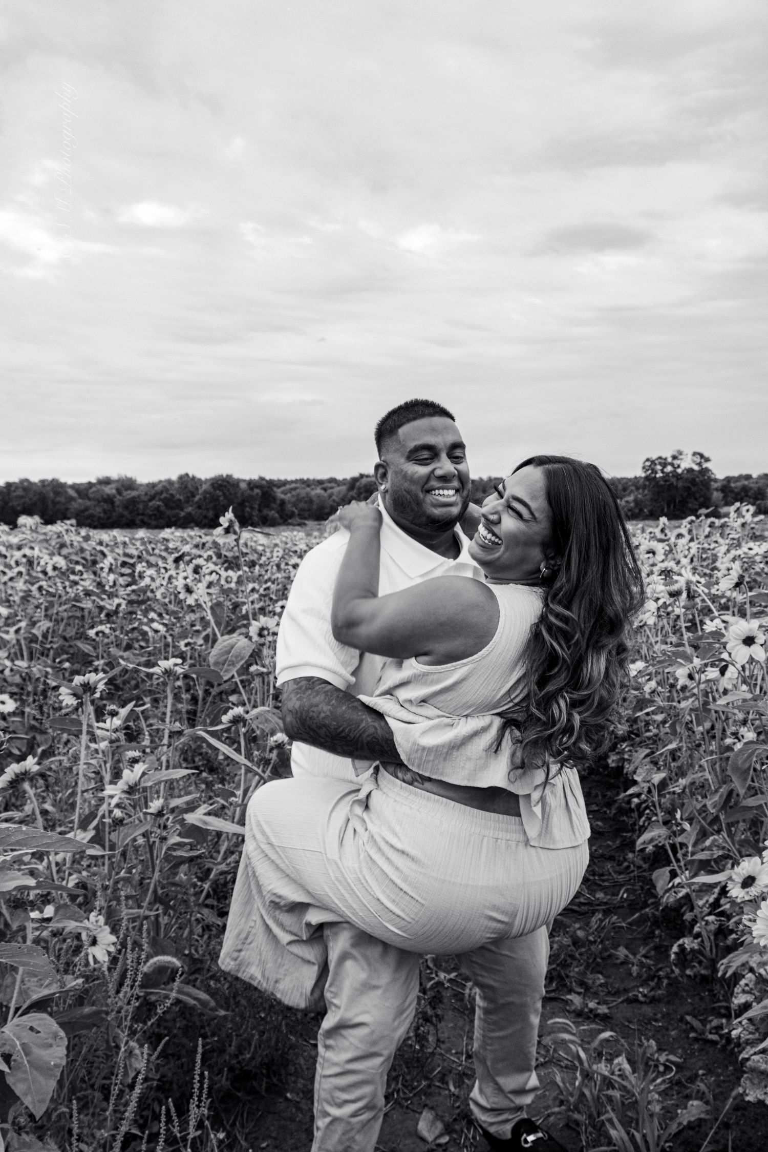 Black and white photo of a couple embracing and laughing together in a sunflower field on a cloudy day.