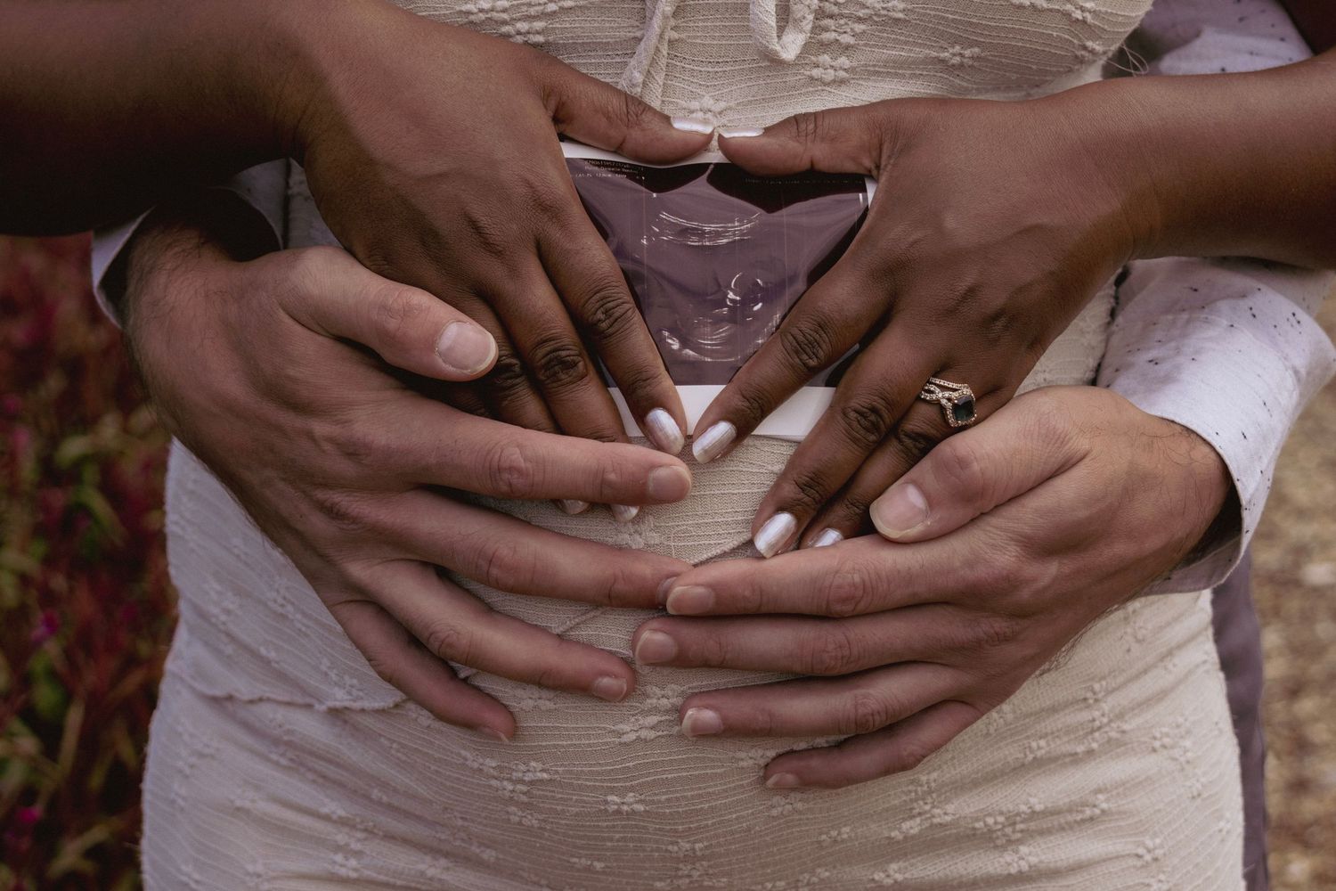 Couple forming heart shape with hands on pregnant belly, wearing wedding rings, maternity photo.