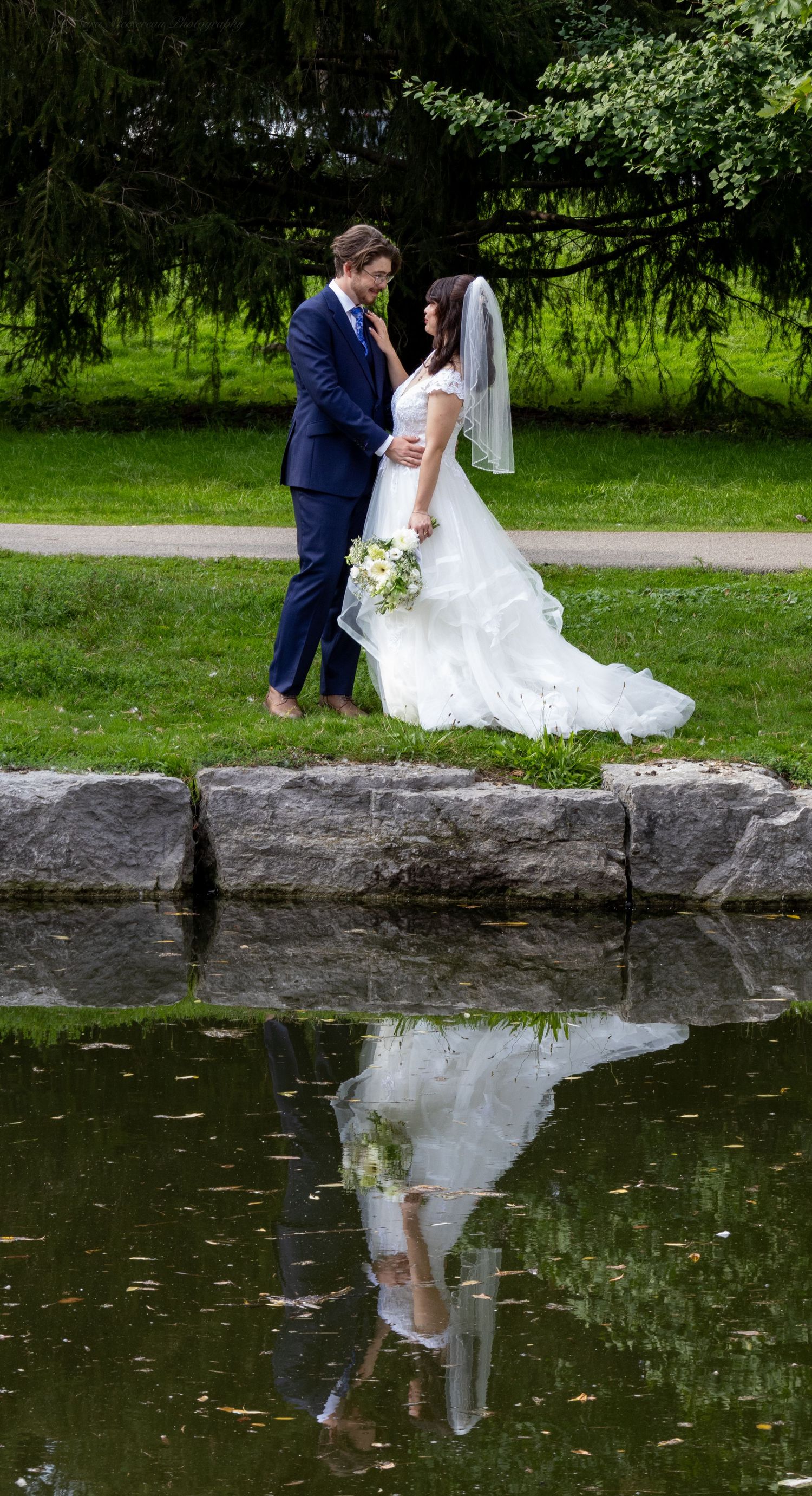 Bride and groom share a romantic moment by a reflective pond in a lush green park, her white gown and bouquet mirrored in the water.