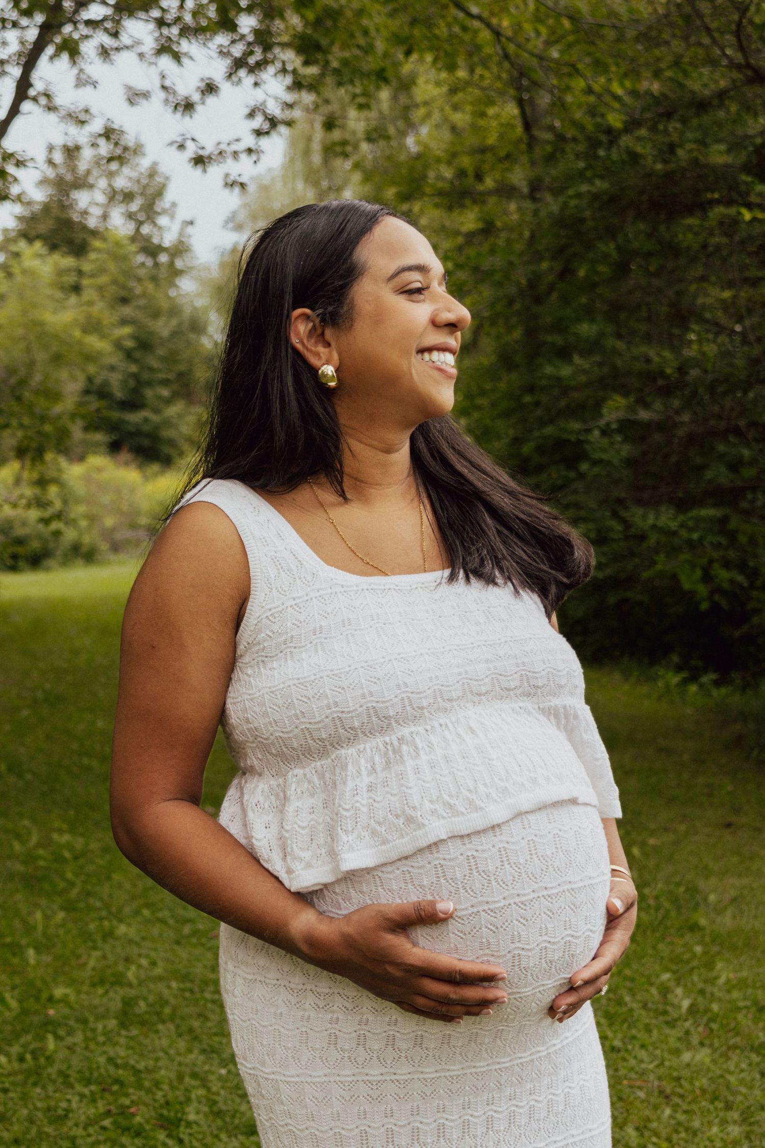 Happy pregnant woman in white dress cradling her baby bump while standing in a green outdoor setting.