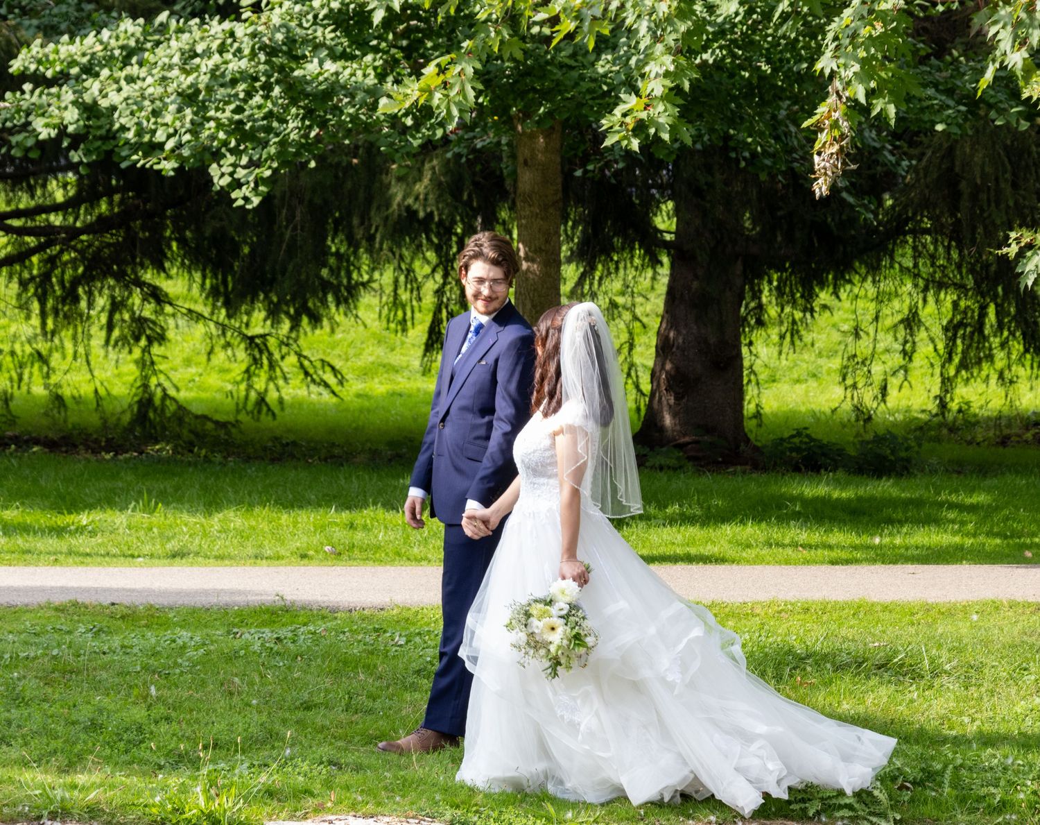 Bride in white gown and groom in navy suit walk hand in hand through a lush green park on their wedding day.