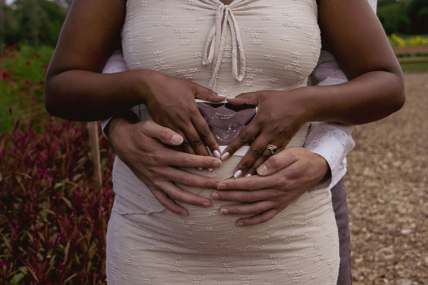 Pregnant woman in white dress with partner's hands forming heart shape on baby bump outdoors.