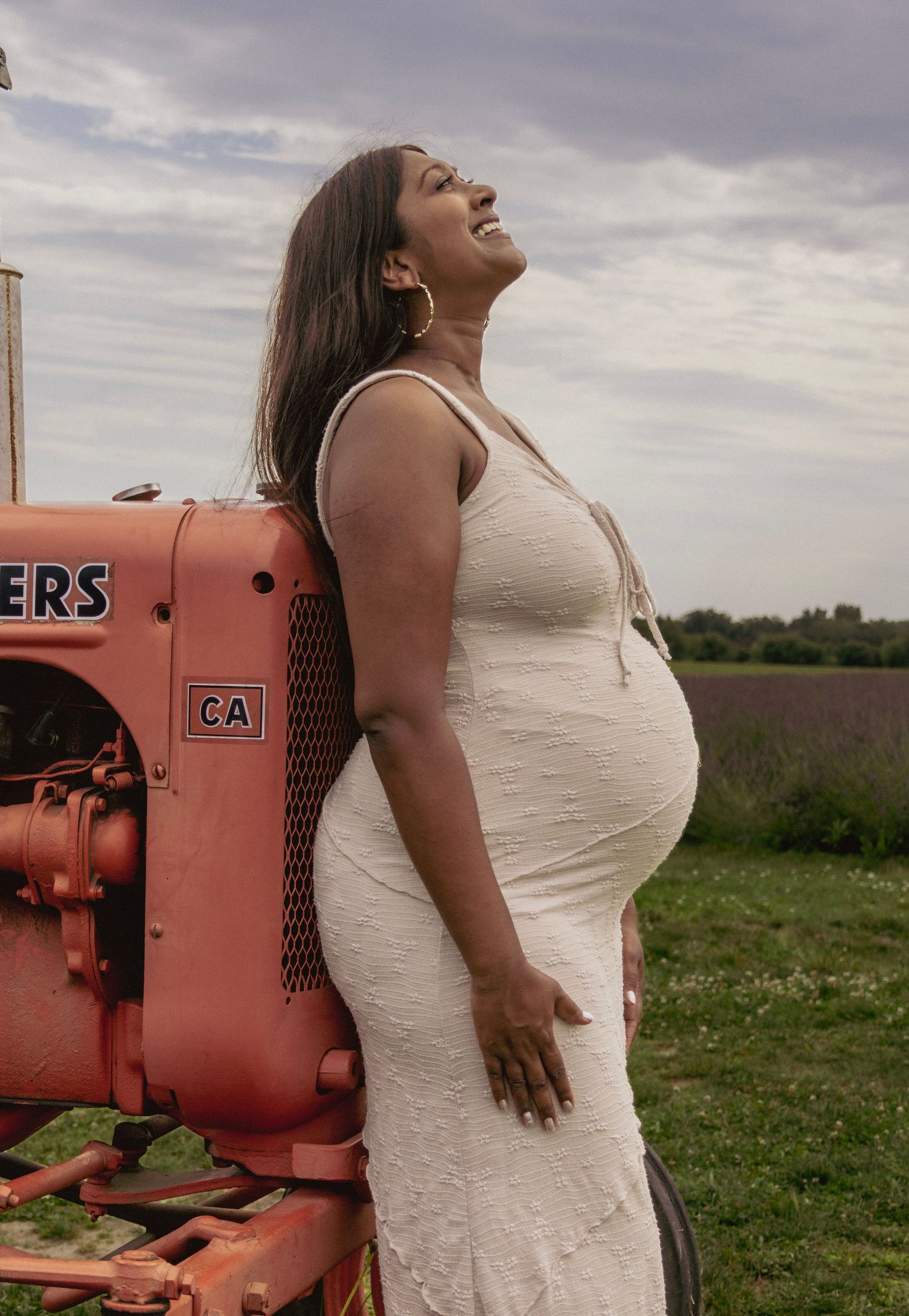 Pregnant woman in white dress leaning against vintage red tractor in a farm field, smiling upward.