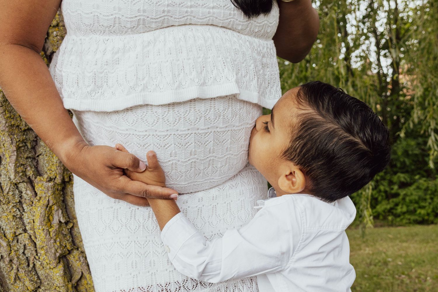Young boy kissing pregnant mother's belly outdoors, both wearing white clothing near a tree.