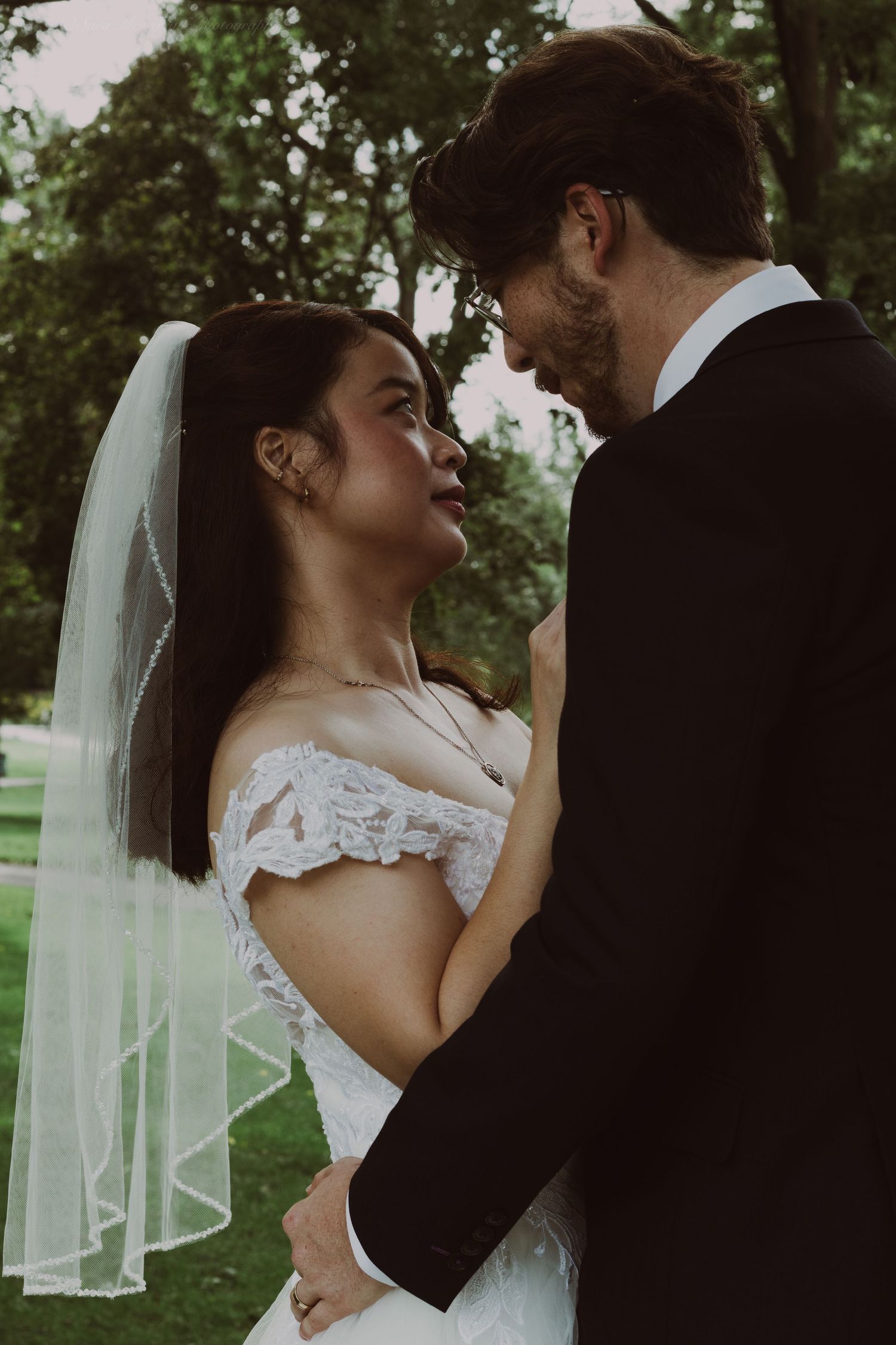 A bride in a white lace off-shoulder gown and veil gazes lovingly at her groom in a black suit outdoors.