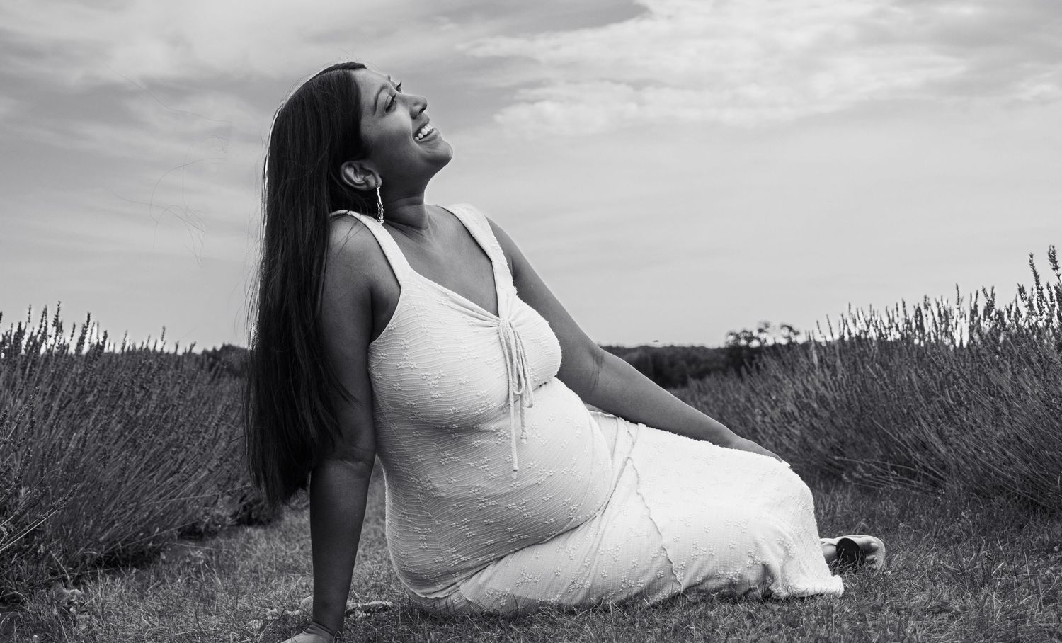 Pregnant woman in white dress sitting in lavender field, head tilted back, long dark hair, black and white photo.