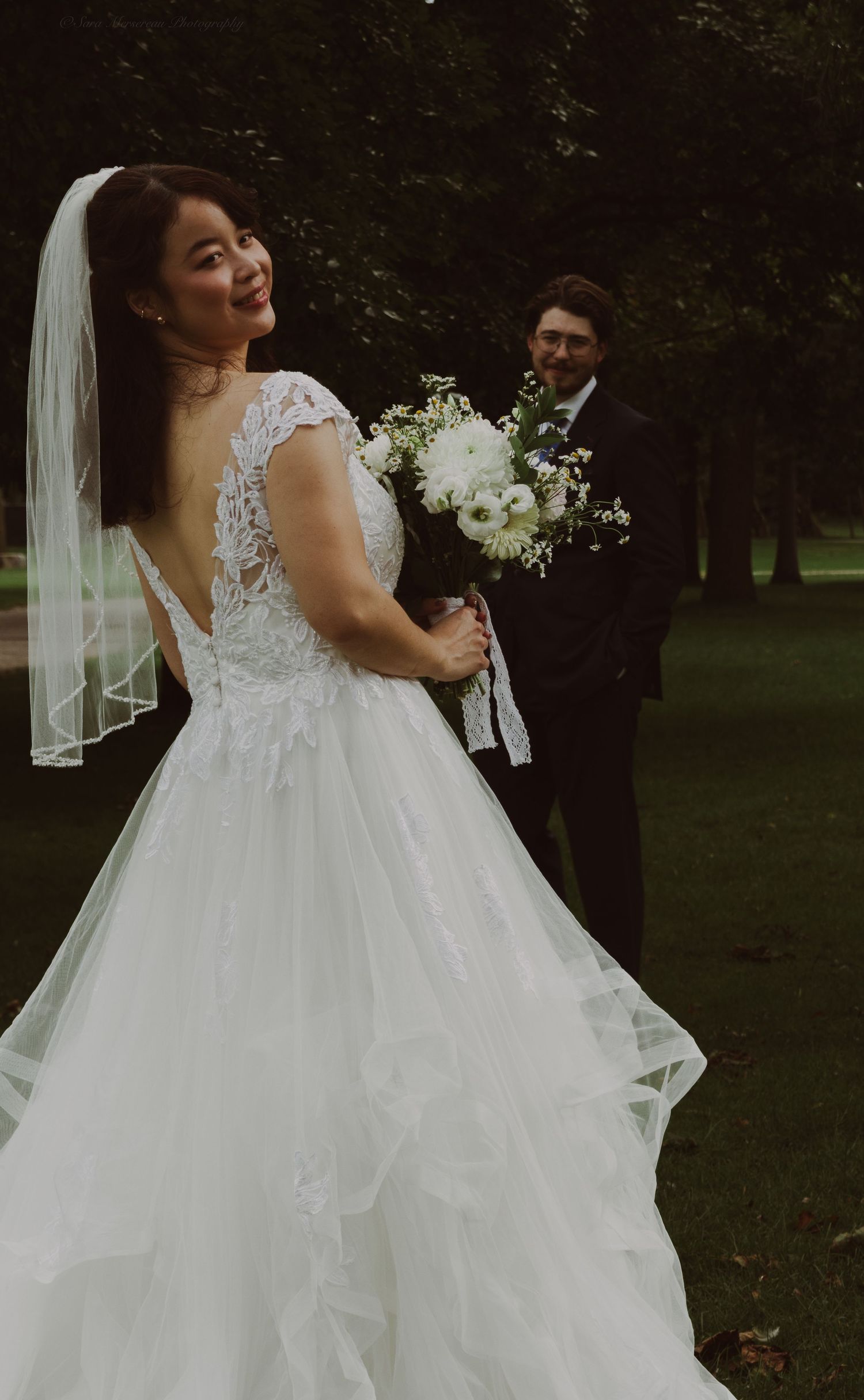 Bride smiles over shoulder showing lace back of white ballgown with veil, groom in black suit behind her.