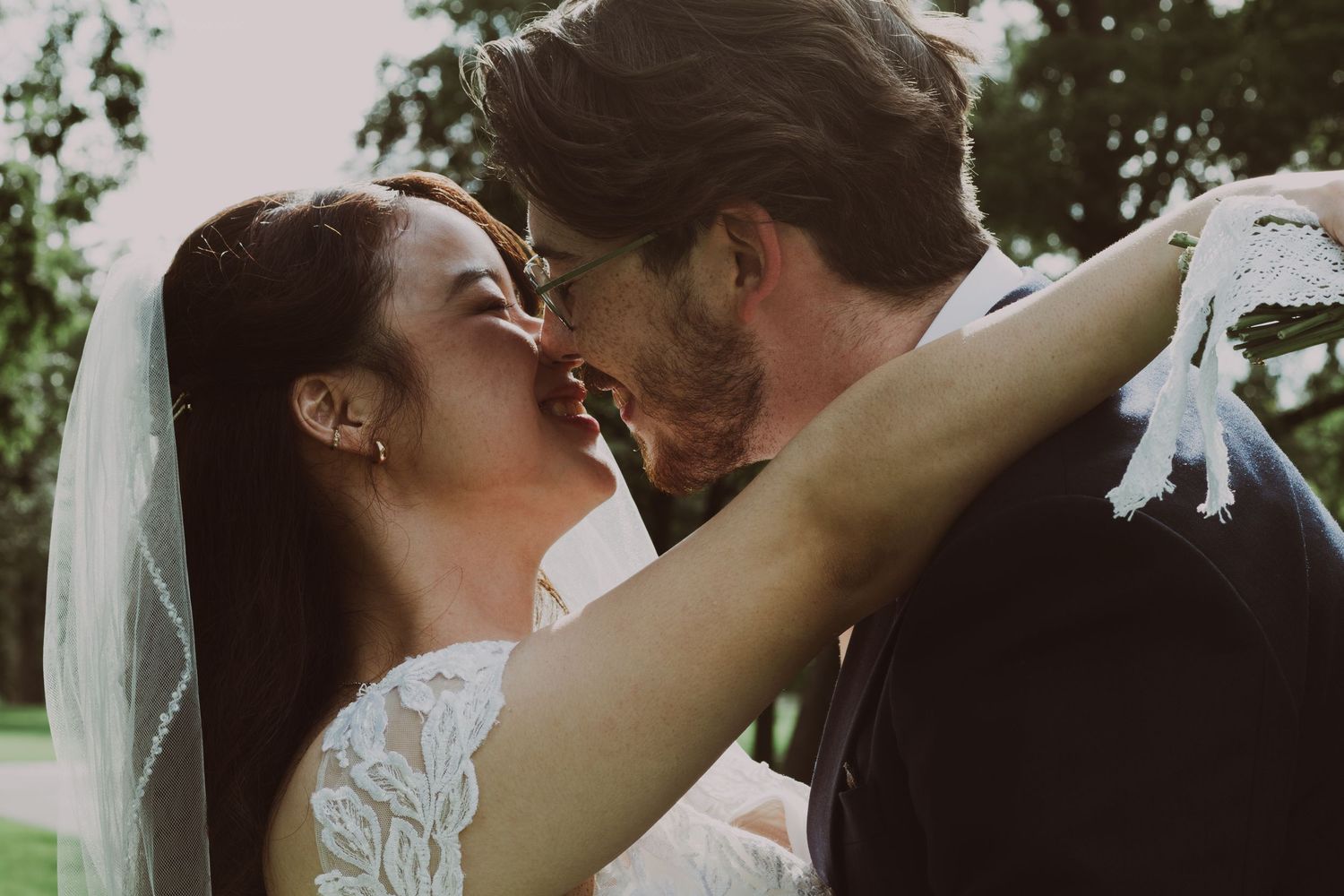 Bride and groom sharing a romantic kiss outdoors on their wedding day, with bride wearing lace gown and veil.