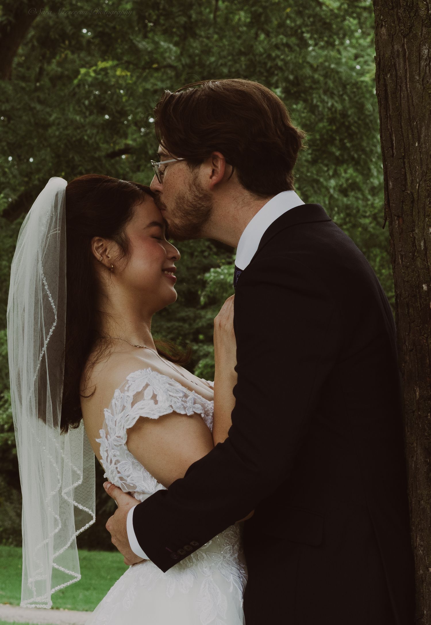 A groom in a black suit kisses his bride wearing a lace dress and veil outdoors among green trees.