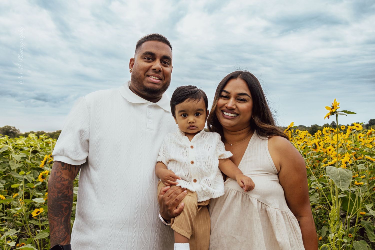 Happy family of three posing together in a beautiful sunflower field on a partly cloudy day.