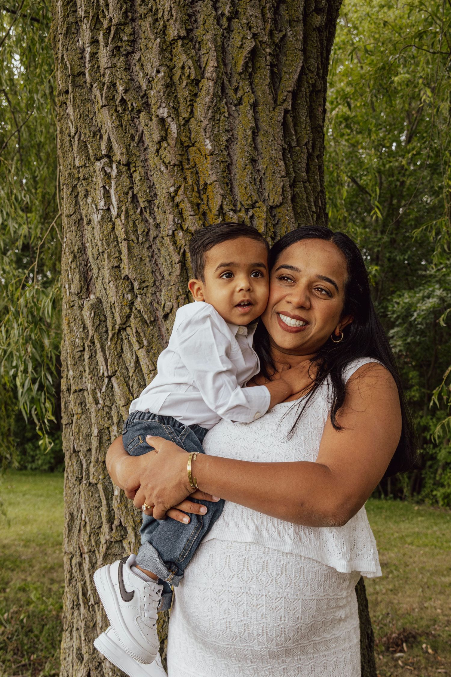 A smiling woman in a white dress holds a young boy near a tree in a lush green park.