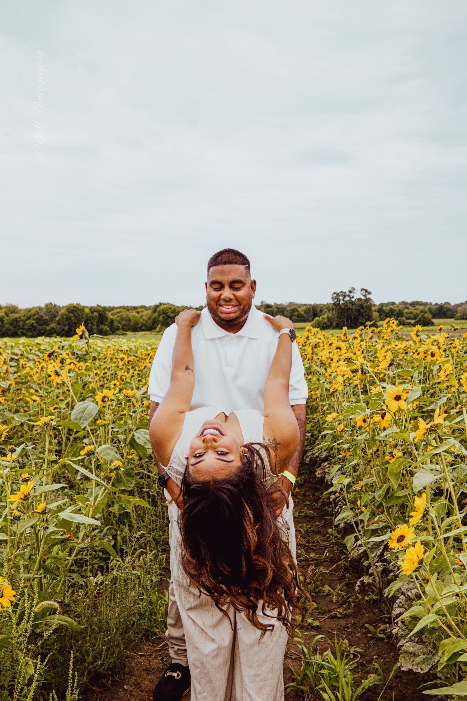 Man joyfully lifting woman upside down while laughing together in a vibrant yellow sunflower field.
