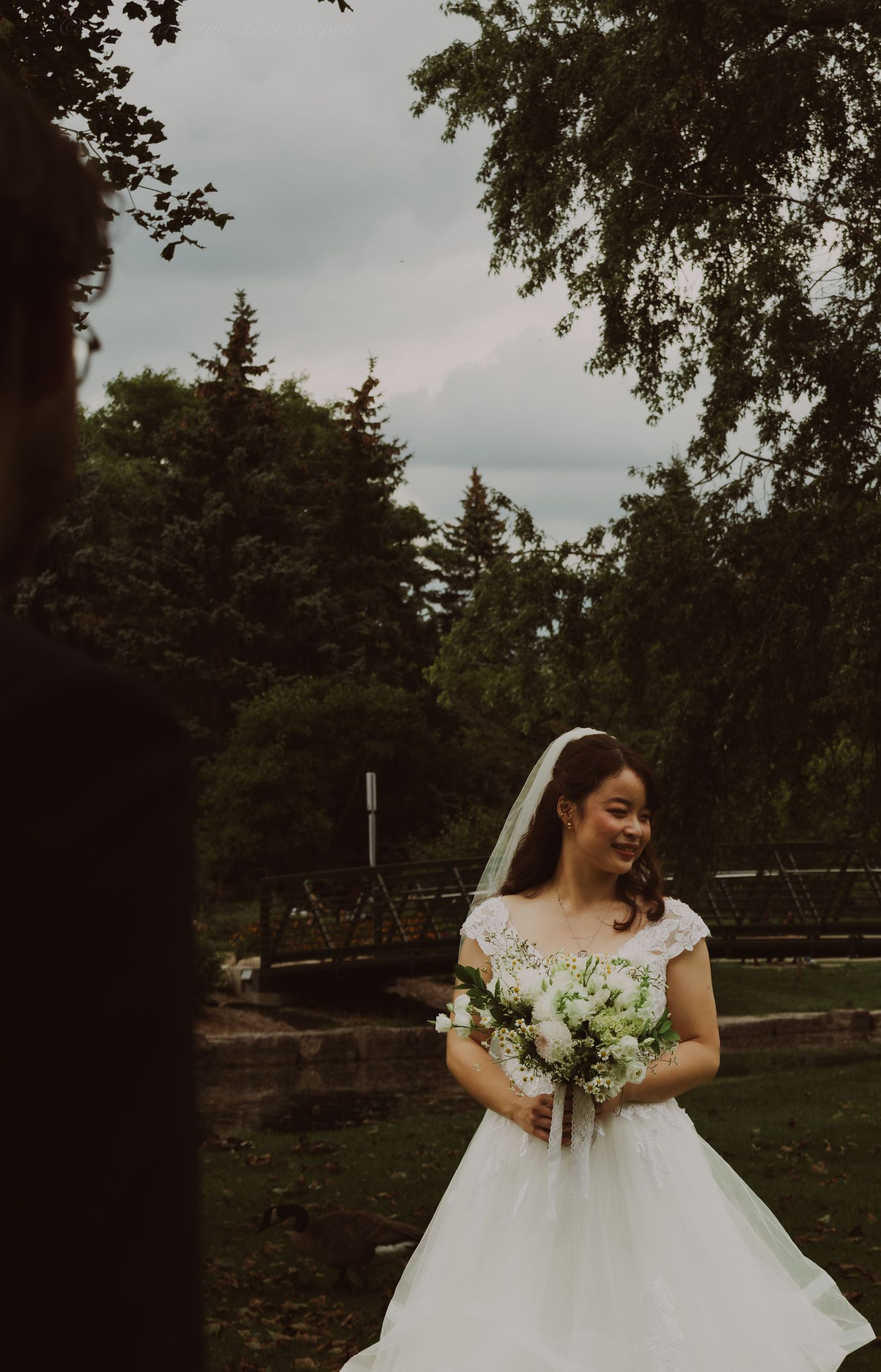 Smiling Asian bride in white lace gown and veil holds bouquet outdoors near bridge surrounded by lush green trees.