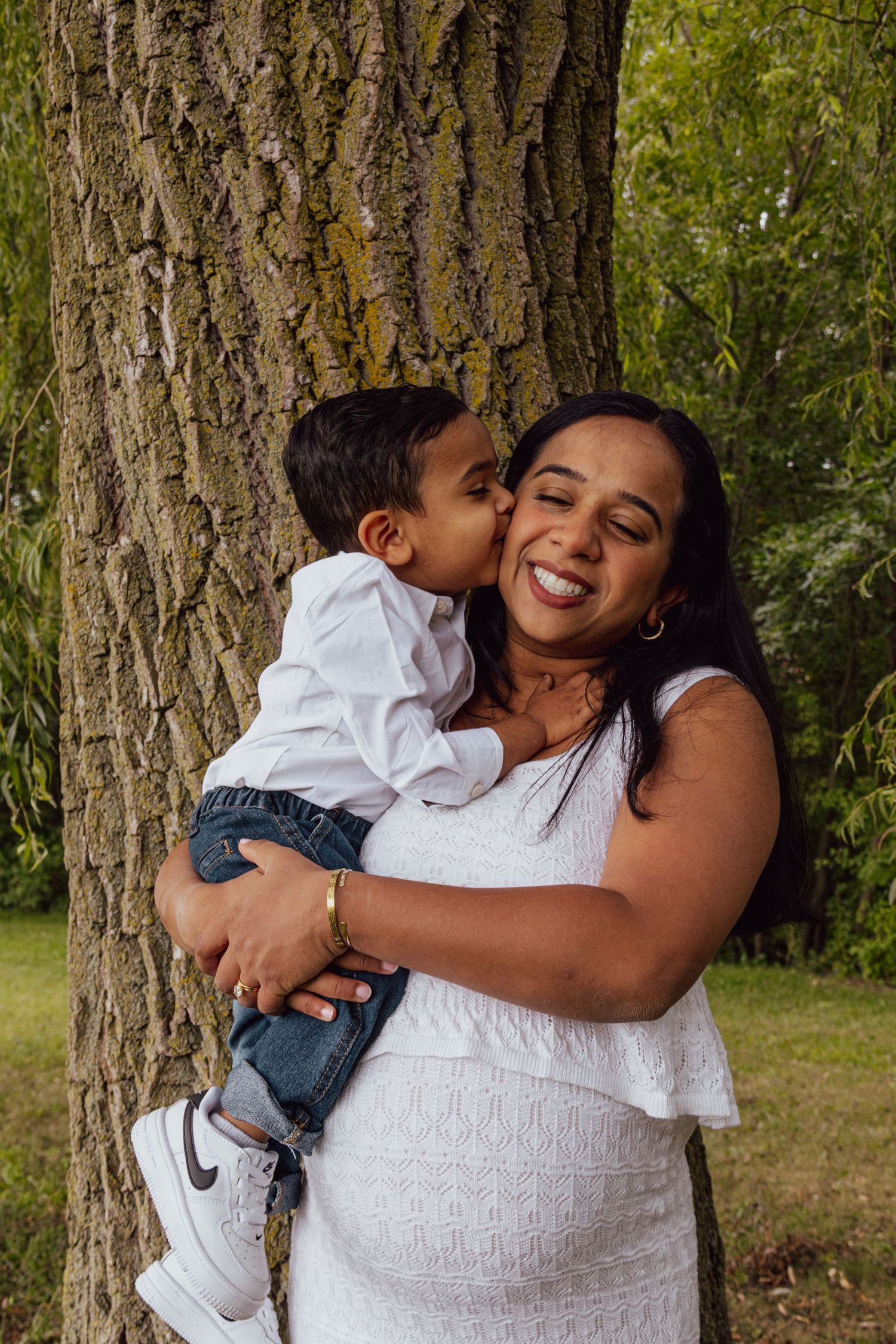 Smiling pregnant woman in white dress holds toddler son who kisses her cheek by a tree.