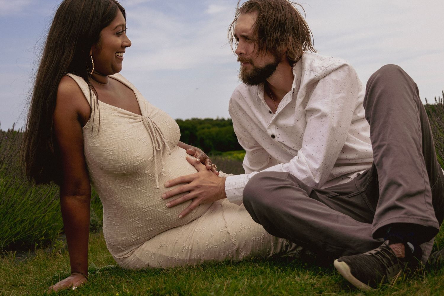 Pregnant woman and bearded man sitting on grass, sharing a tender moment outdoors in a lavender field.