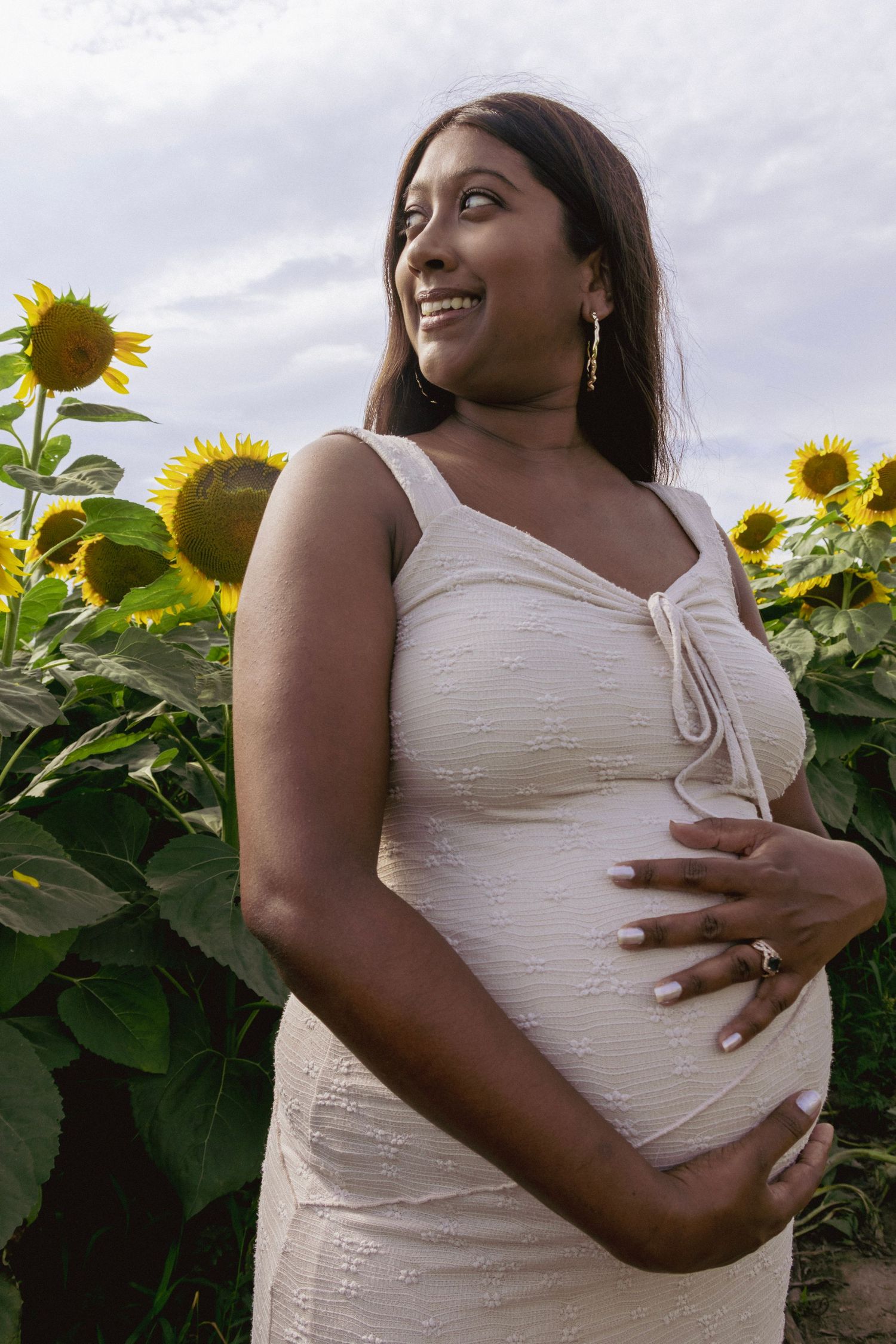 Smiling pregnant woman in white dress holding belly in sunflower field.