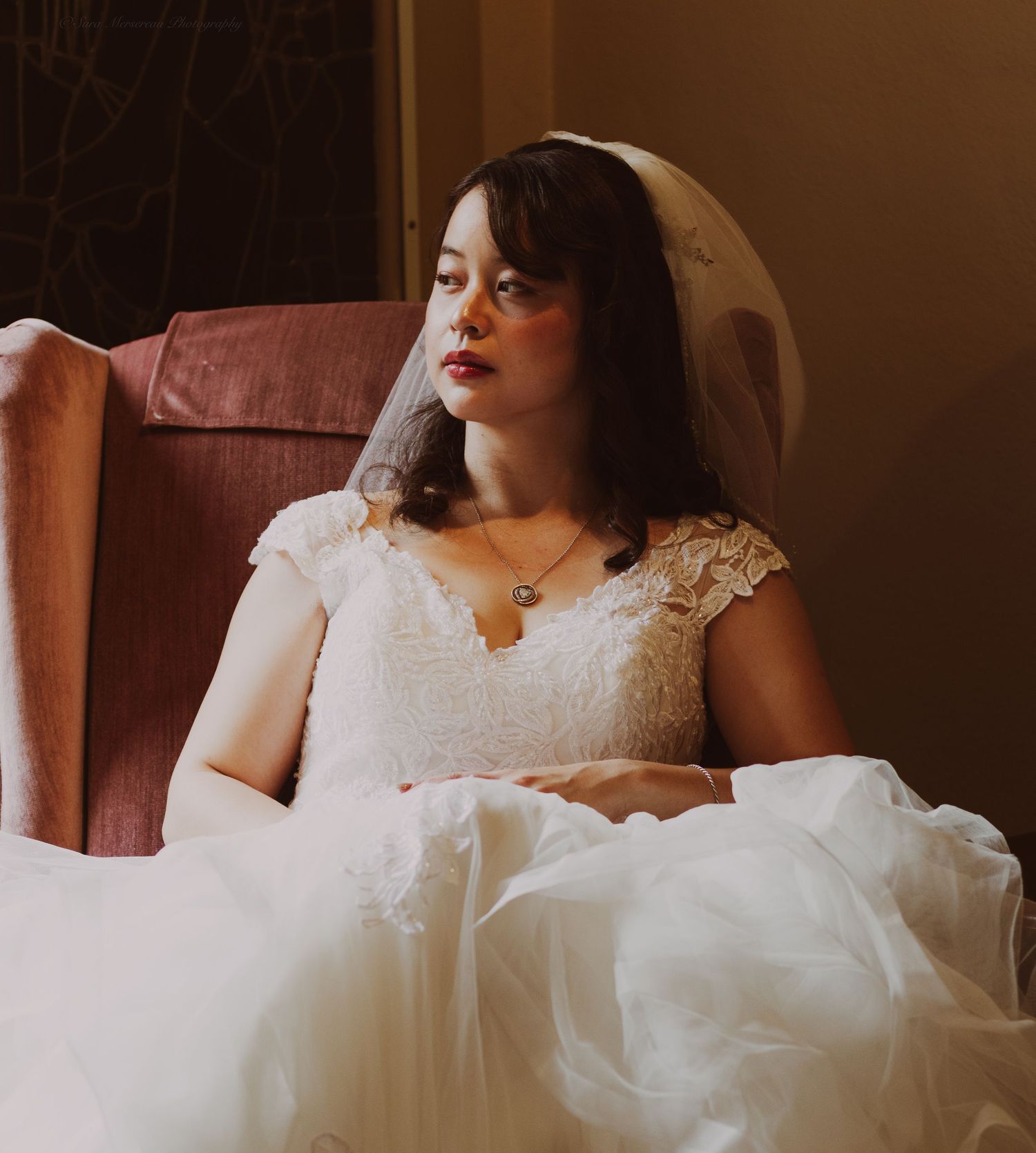 A bride in a white lace wedding dress and veil sits elegantly in a velvet chair, gazing thoughtfully to the side.