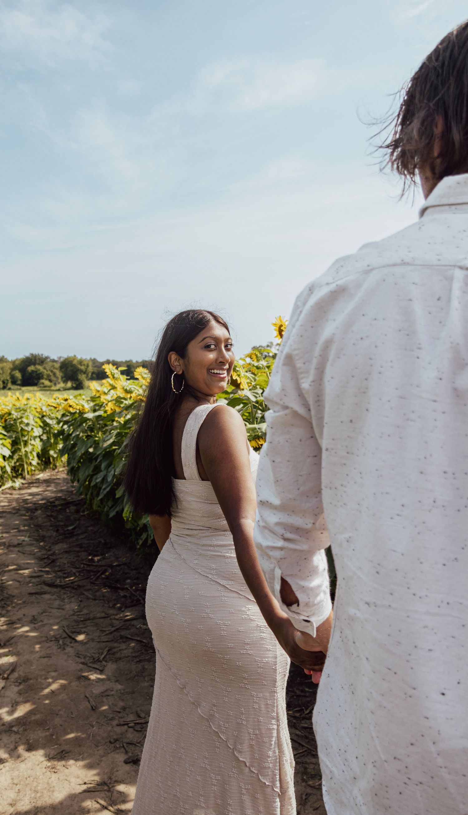 Couple holding hands in a sunflower field, woman in white dress smiling over her shoulder on a sunny day.