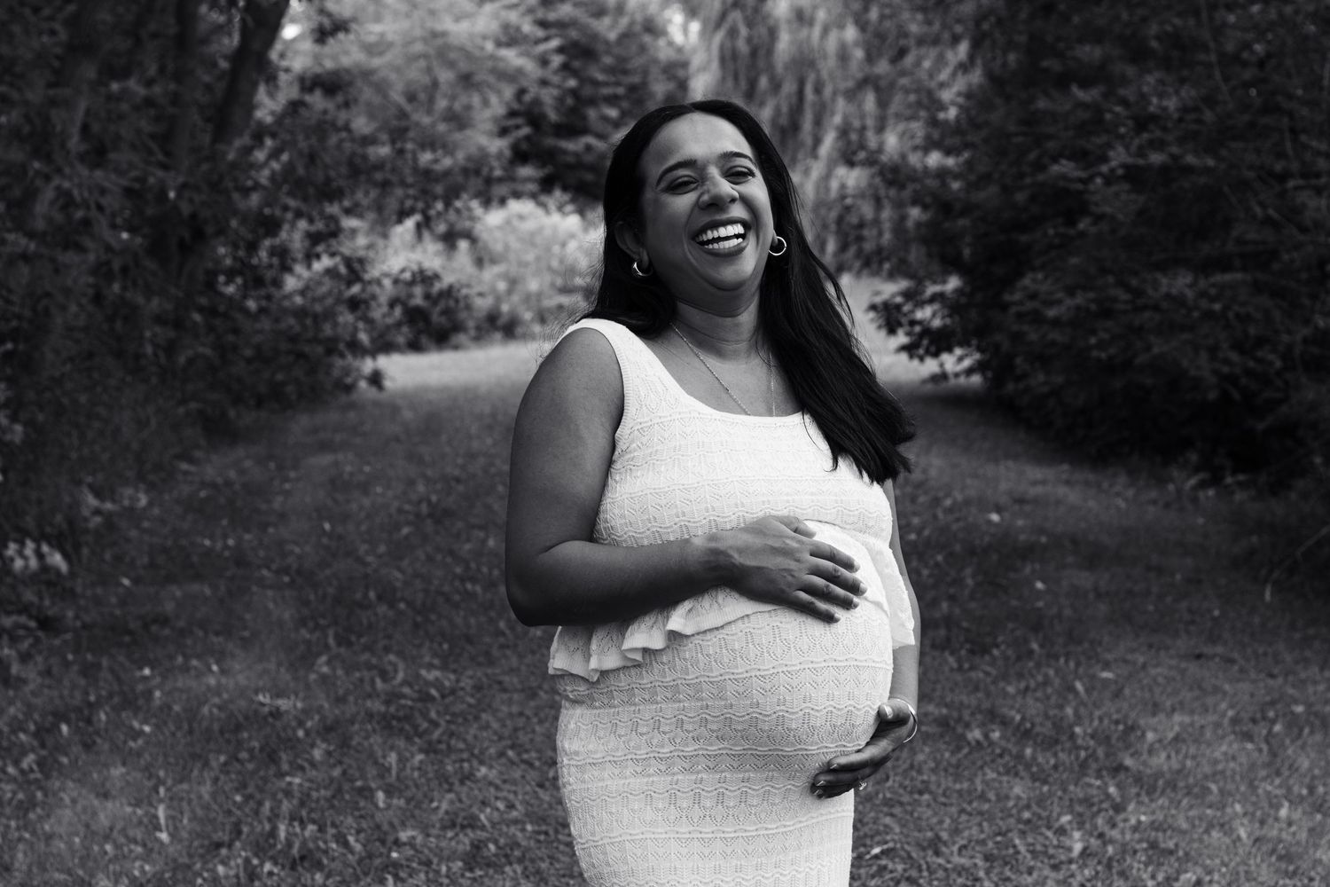 Smiling pregnant woman in white dress holding baby bump outdoors in black and white maternity photo.