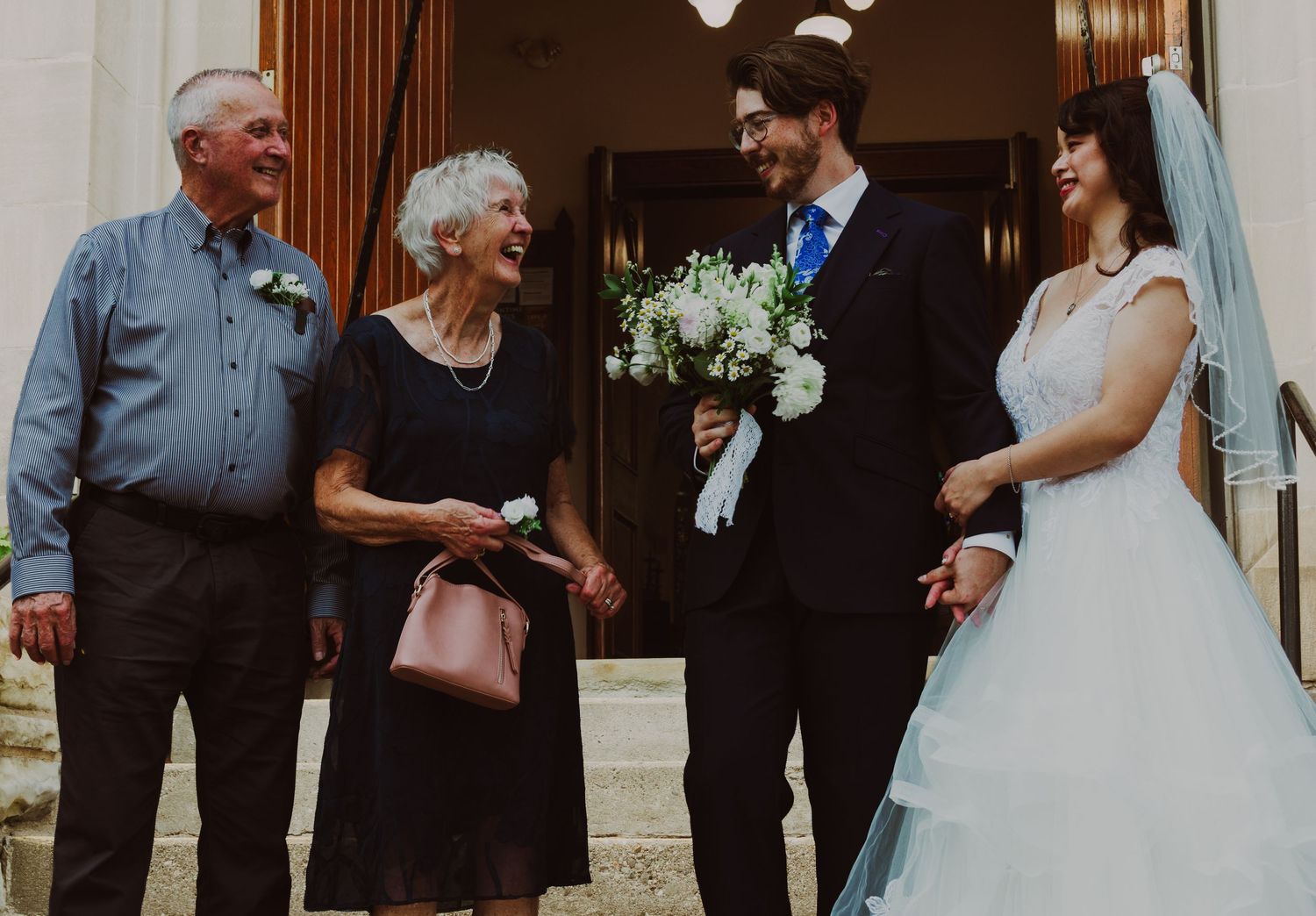 Bride and groom smiling with elderly couple outside a historic building on their wedding day.