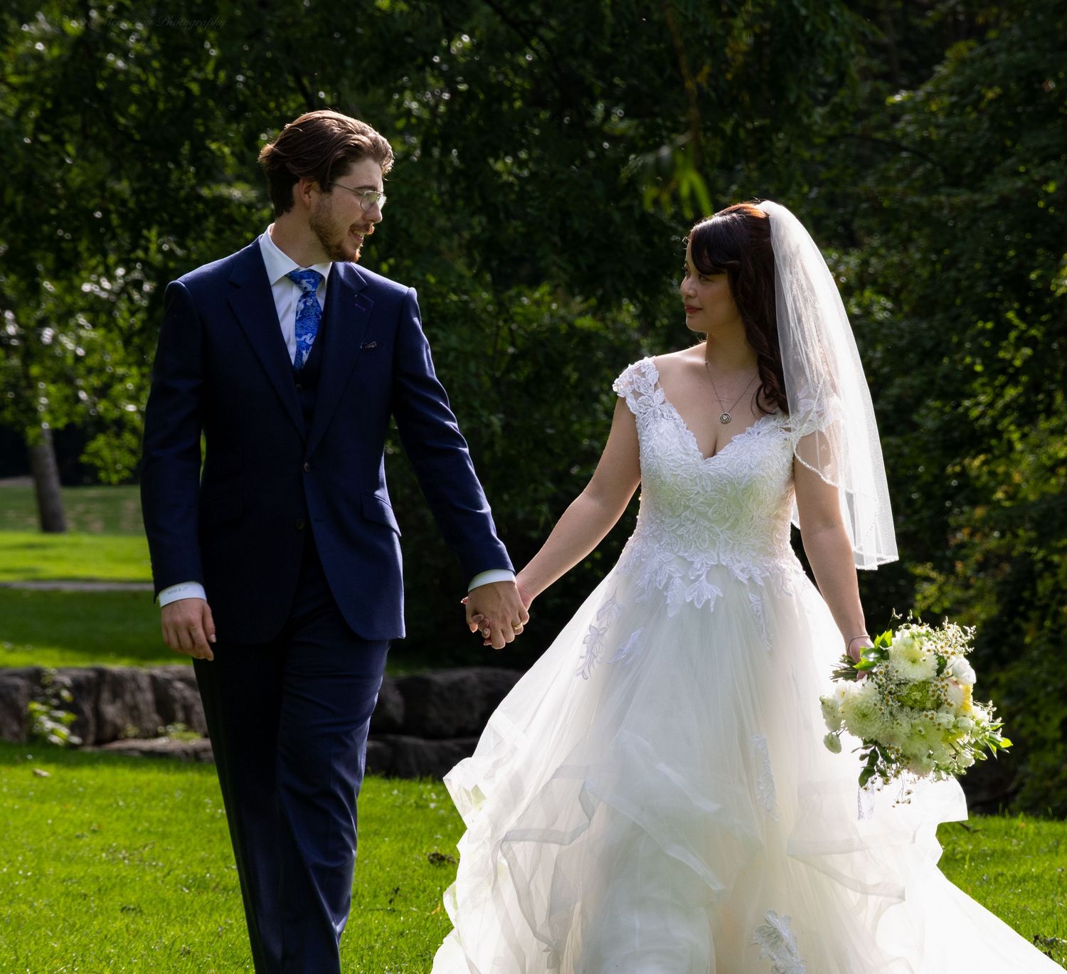 Bride in white gown holding bouquet walks hand in hand with groom in navy suit on green lawn.