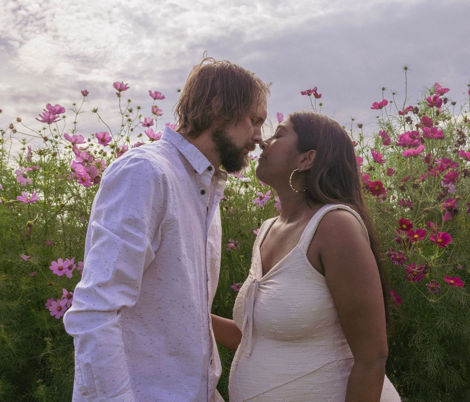 Couple sharing a romantic kiss in a vibrant flower field with pink cosmos blooms under a cloudy sky.