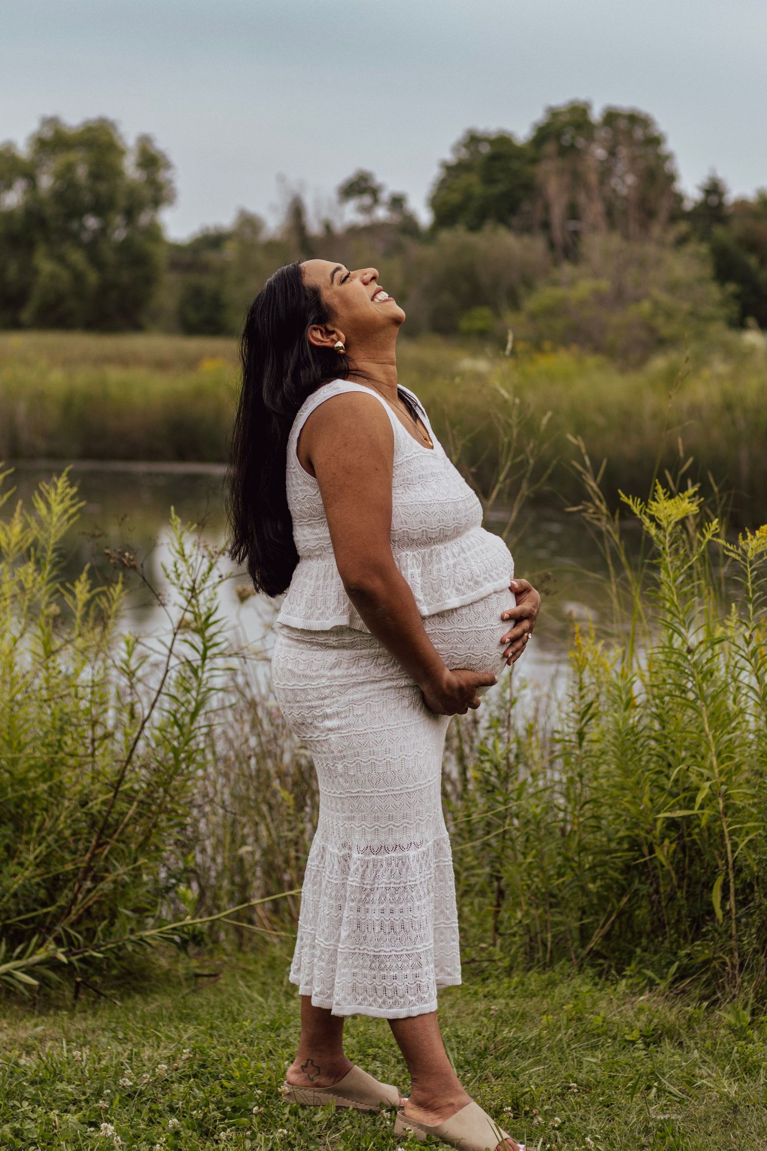 Pregnant woman in white lace dress laughing joyfully outdoors near a pond, cradling her baby bump.