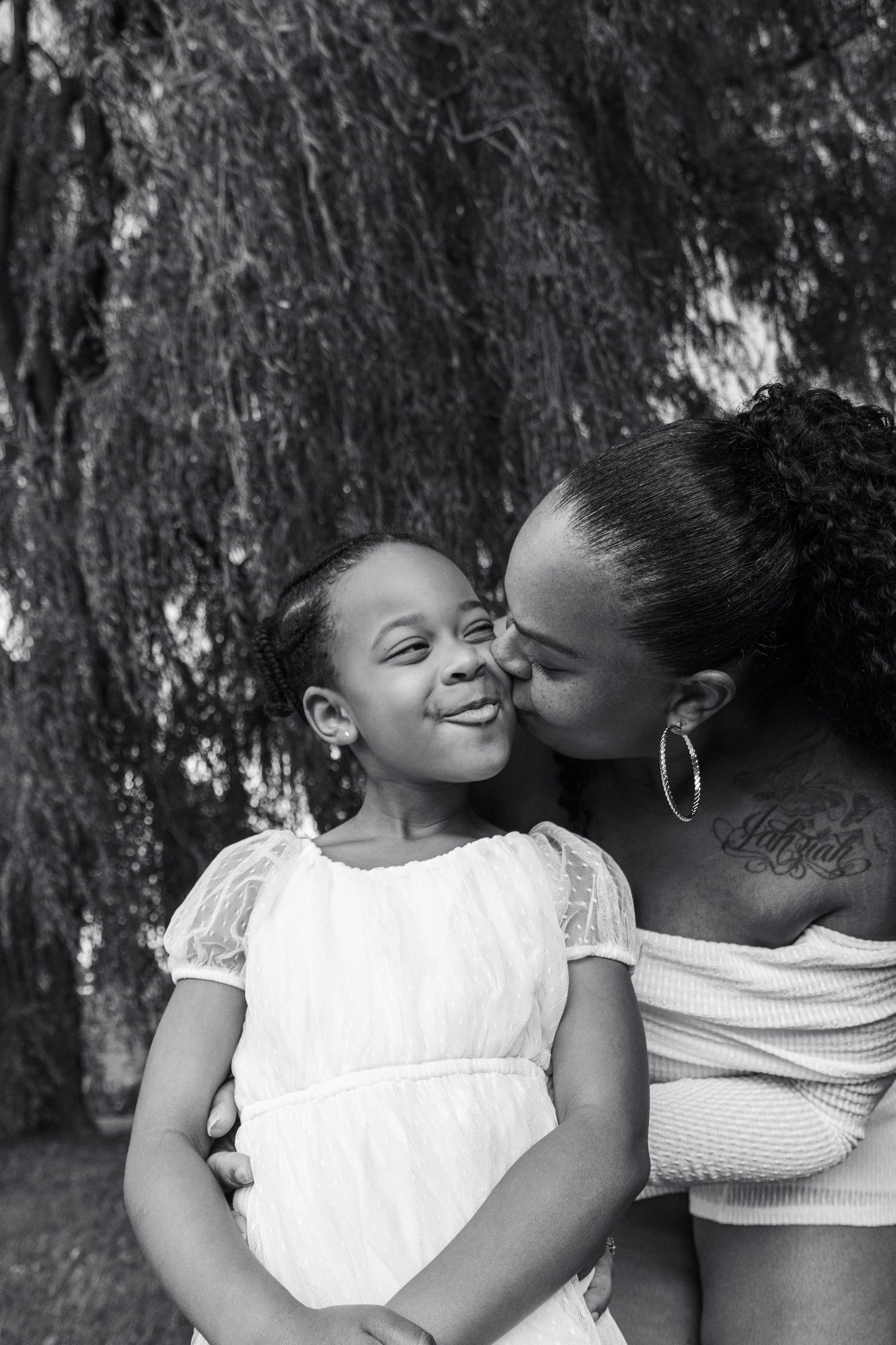 A mother tenderly kisses her smiling young daughter on the cheek in a black and white outdoor portrait.