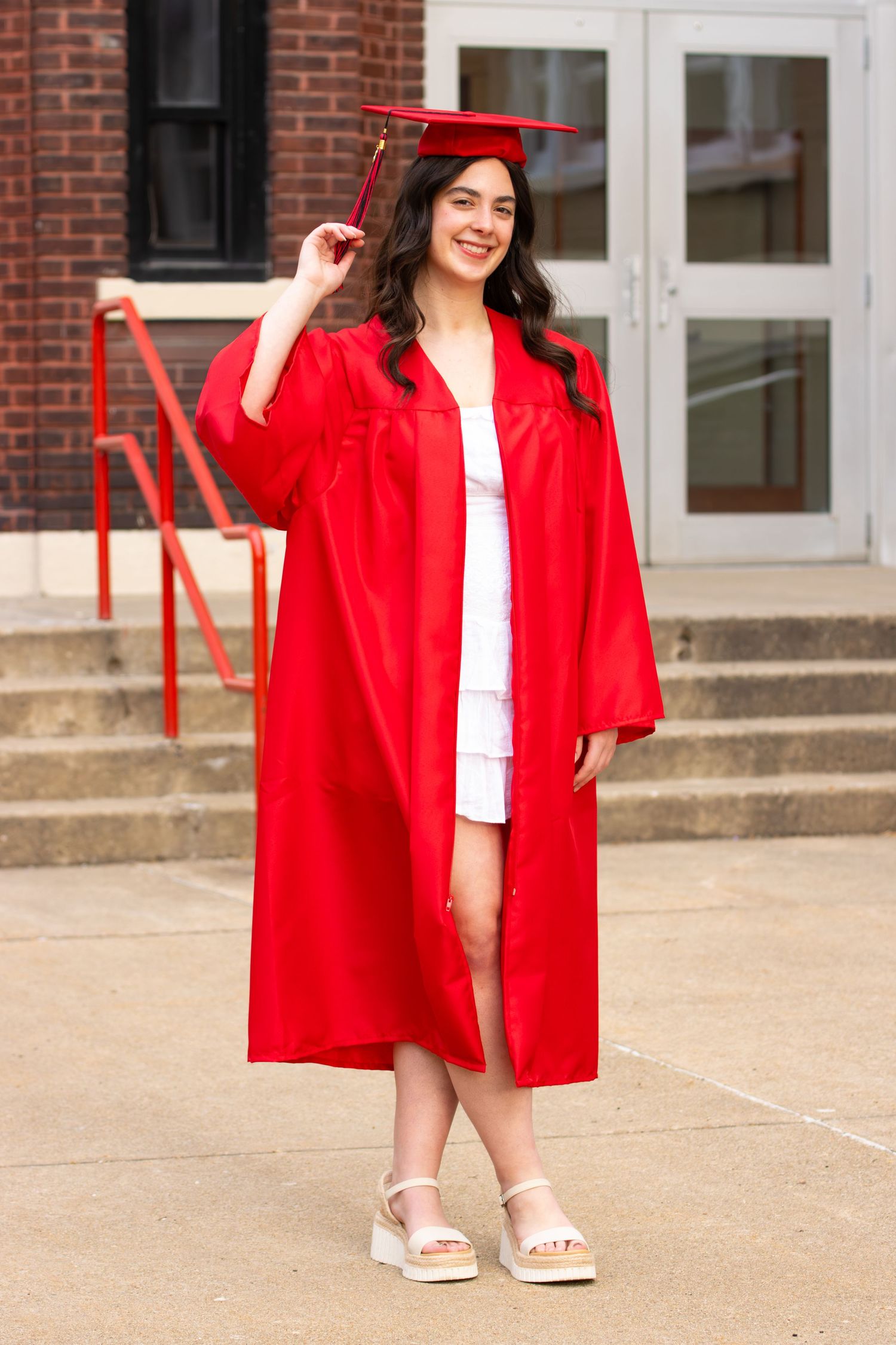 Young woman in red graduation cap and gown smiling outdoors on school steps.