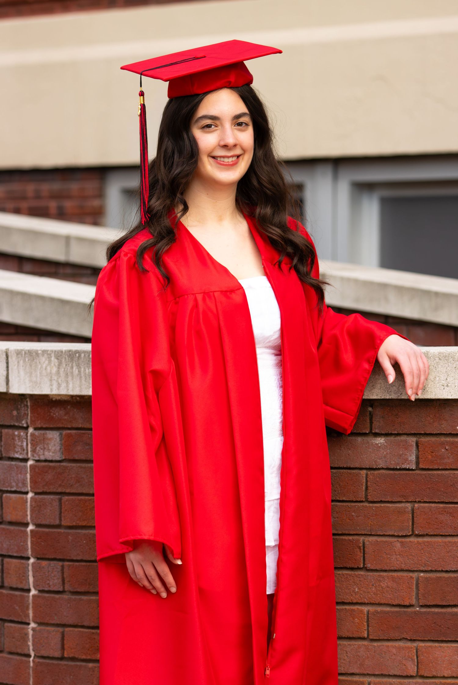 Smiling graduate in red cap and gown posing against brick wall outdoors.